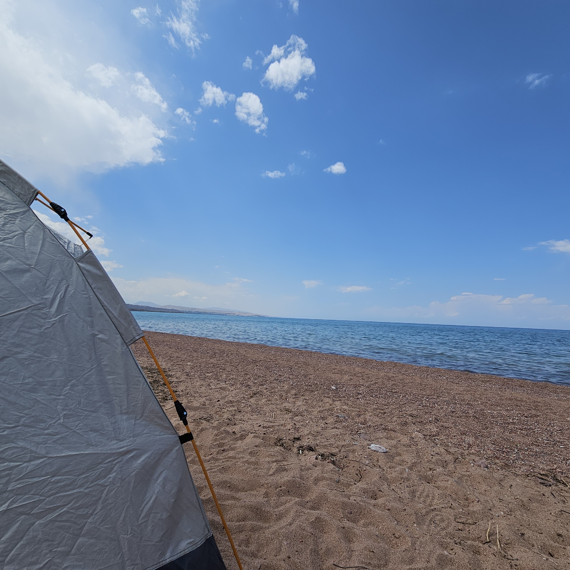 An eye-level, wide-angle shot captures a serene view of a beach in Kaji-Say, Kyrgyzstan. The foreground is dominated by the partial view of a grey tent, its orange poles and black clips visible against the sand. The beach itself is composed of fine sand and small pebbles, with scattered dry grass and a piece of white plastic debris visible. Beyond the beach, the calm, blue waters of Issyk-Kul lake stretch towards the horizon, meeting a distant, hazy shoreline with hills. The sky above is a vibrant blue, dotted with scattered white cumulus clouds. The overall impression is one of tranquility and natural beauty, with the tent suggesting a moment of outdoor recreation or camping.
