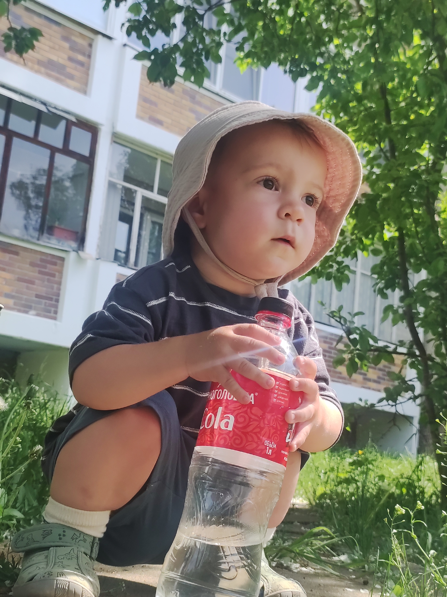 A young child in Chaikovsky, Russia, wearing a light-colored hat, striped shirt, and blue shorts, crouches outdoors, holding a plastic bottle of red and clear liquid, labeled "Cola" in Russian. The child wears white socks and green sandals. Behind the child is a building with windows and a brick facade, partially obscured by tree branches. The scene is in daylight.