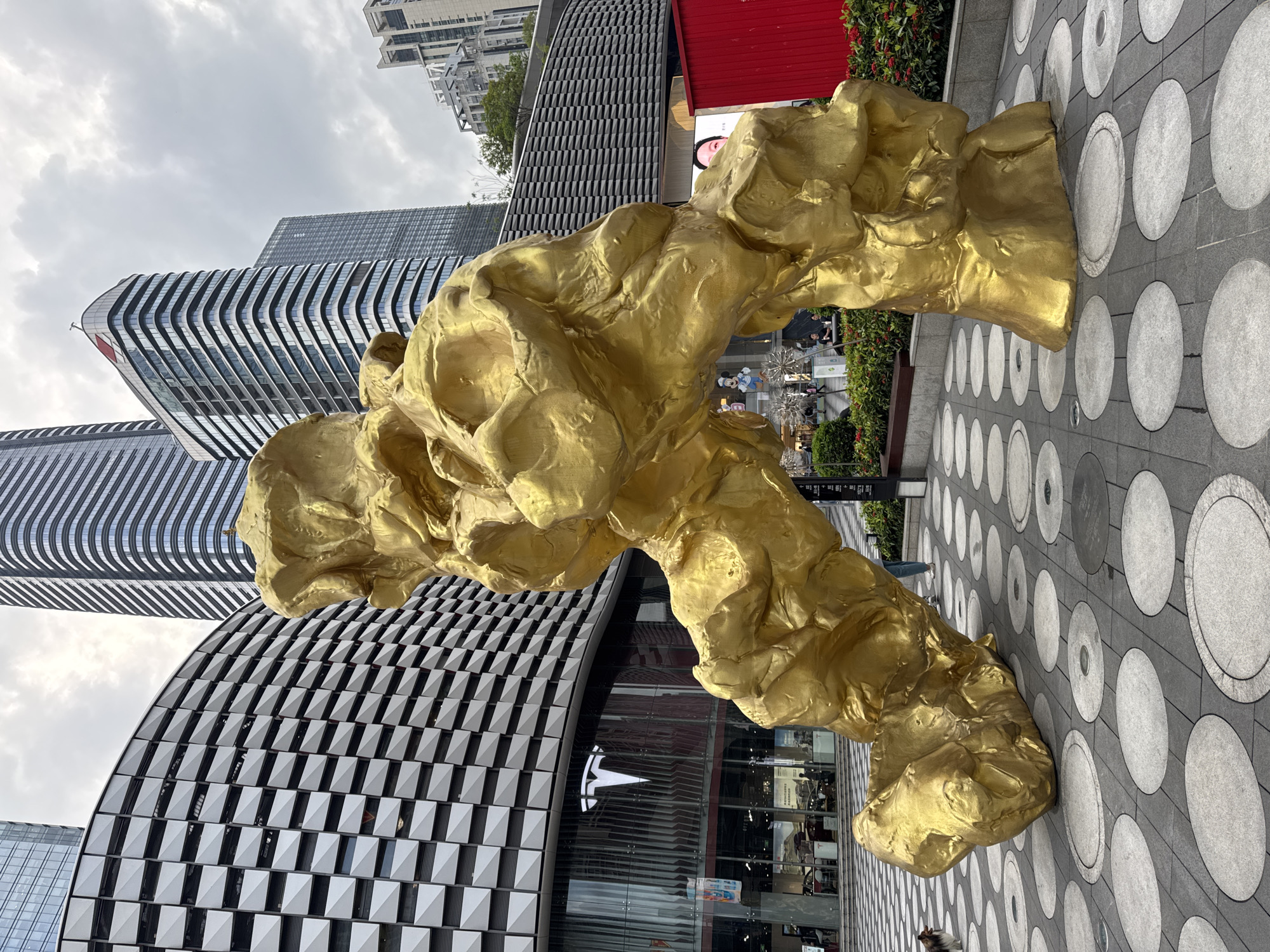 The image shows a large, golden, abstract sculpture in an urban setting. The sculpture is positioned on a gray paved surface with large, circular patterns. The background features modern architecture, including tall, geometric buildings. A Tesla store is visible, indicated by the logo. People are present, though not the main focus. The location is identified as Futian District, China, based on the style of architecture and urban design.