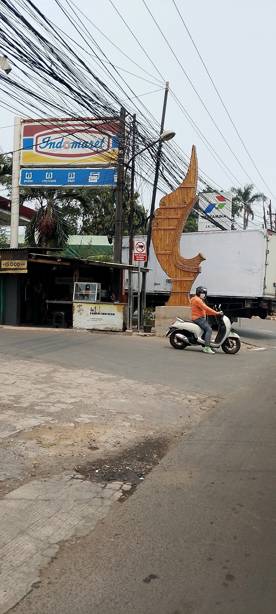 The image captures a street scene in Cileungsi, Indonesia, dominated by a dense network of overhead electrical wires. In the foreground, a person wearing an orange jacket, blue jeans, a helmet, and a face mask rides a white scooter. The scooter is in motion, turning right. To the left of the road, a small food stall with a sign reading "WARUNG TEMI IS FRIED CHICKEN" is visible. Above the stall, a large "Indomaret" sign is displayed. Further back, a white truck is parked, and behind it, a large, decorative bamboo structure stands. Another sign for "Pertamina" is also visible in the background. The road itself appears to be asphalt, with some areas of cracked pavement and a small puddle. The sky is overcast.
