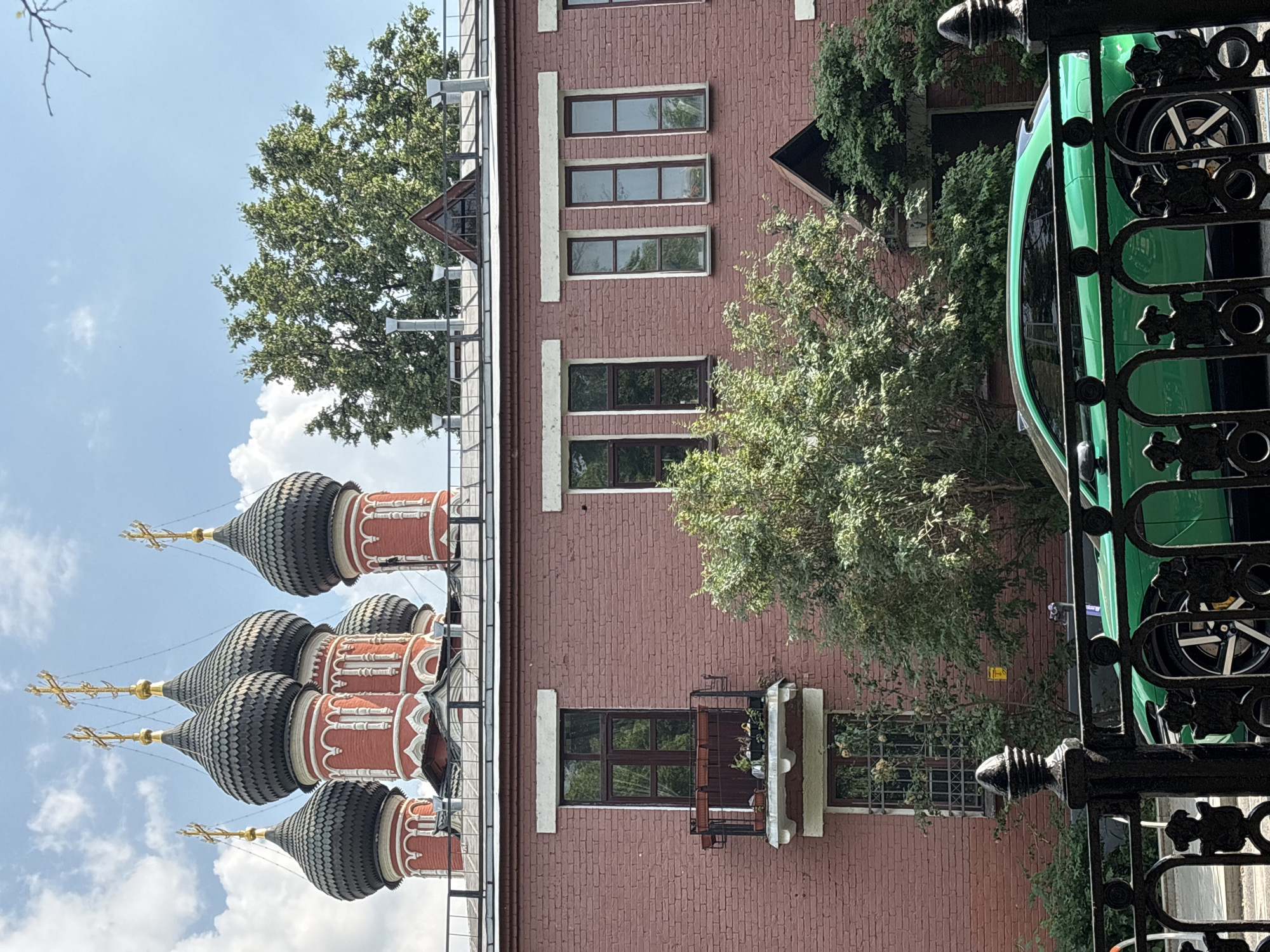 The image shows the Church of the Resurrection of Christ in the Kadashi district of Moscow, Russia. The church's distinctive red brick facade and onion domes are visible behind a modern brick building with several windows and a small balcony. In the foreground, a bright green sports car is parked behind a black wrought-iron fence. The sky is blue with scattered clouds. The overall scene depicts a juxtaposition of historical and modern architecture in an urban setting.