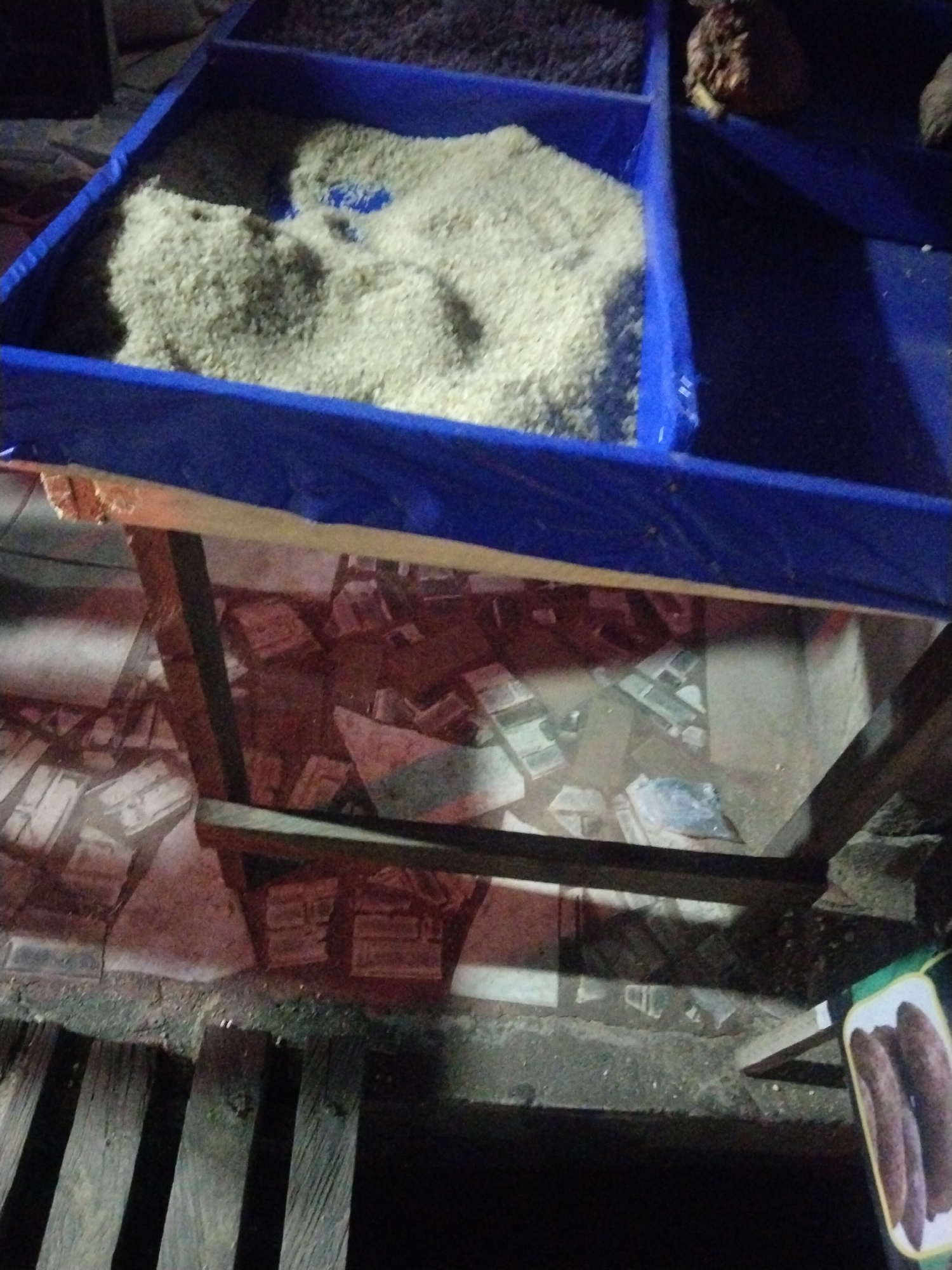 The image shows a market stall in Uyo, Nigeria. The stall is constructed from wood and covered with a blue plastic sheet, forming several compartments containing rice, beans, and possibly dried goods. The floor beneath the stall is tiled in a pattern resembling cracked stone, and adjacent to the stall is a wooden plank walkway. There's a package of dried yam visible at the edge of the image. The overall lighting is poor, suggesting an evening or dimly lit indoor setting. There are no people visible in the frame.