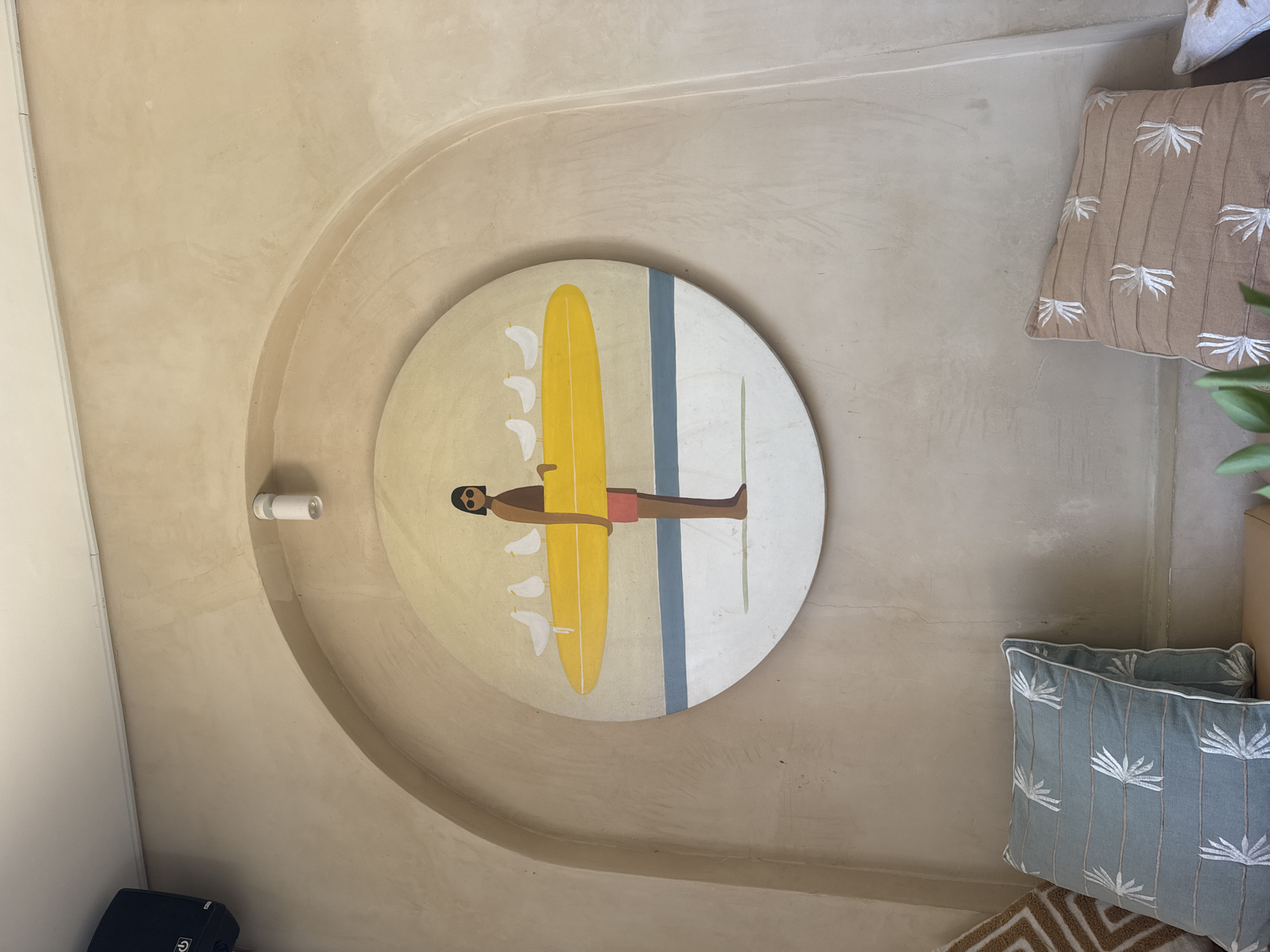 A minimalist painting is displayed on a textured, beige, arched wall in Kuta Selatan, Indonesia. The circular painting depicts a stylized figure holding a yellow surfboard with three white birds perched on it. The background of the painting shows a light yellow sky transitioning to a light blue and white sea and sand. Below the painting, on a bench, are two decorative pillows. One pillow is a pale blue with a white palm leaf pattern, and the other is tan with a similar white palm leaf pattern. A black speaker is mounted on the wall to the left of the painting. A white cylindrical spotlight is positioned above the painting. The wall has a rough, stucco-like texture.