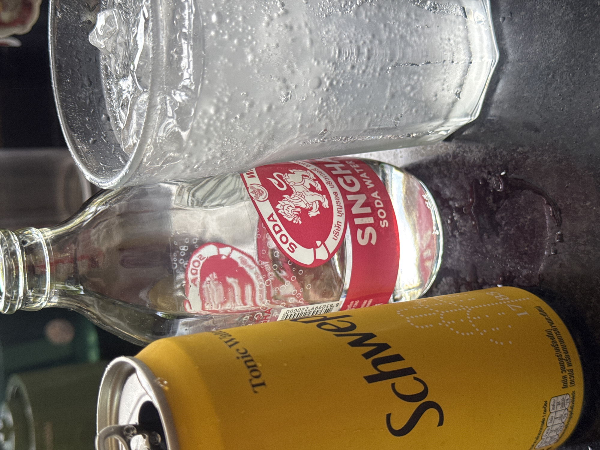 A high-angle, close-up shot shows a yellow Schweppes Tonic Water can next to a clear glass bottle of Singha Soda Water and a glass of soda water with ice. The Schweppes can is in the foreground on the left, with its pull-tab visible. The Singha bottle is in the middle, with a red label featuring a lion. The glass of soda water with ice is on the right. The surface below the items appears to be a dark, speckled countertop with some condensation or spilled liquid. The lighting is bright and casts shadows.