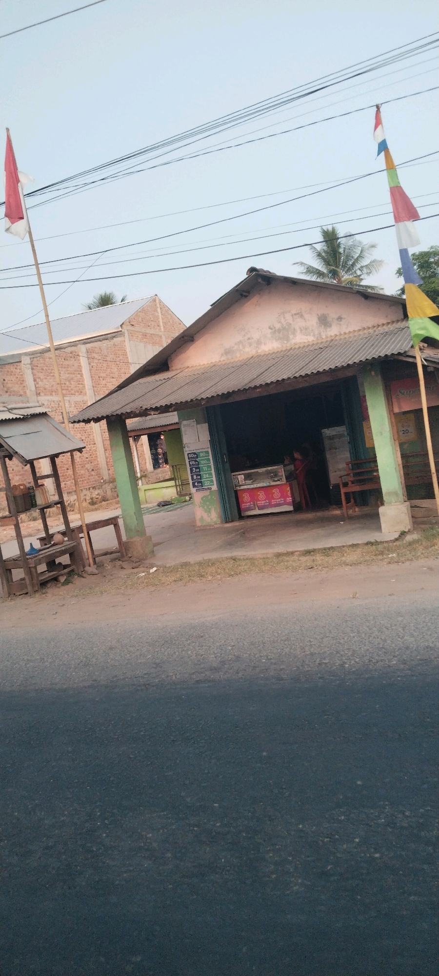 This is a vertically oriented, outdoor, eye-level shot of a roadside shop in Terbanggi Besar, Indonesia. The shop appears to be a small, open-air structure with a corrugated metal roof supported by green painted posts and a weathered, pale pink wall. In front of the shop, a glass display case contains various items, and a person, possibly a cashier or shopkeeper, stands behind it. Two children, a boy and a girl, are inside the shop, seemingly observing the items in the display case. A small wooden bench is placed to the right of the shop entrance. To the left of the shop, a wooden shelf unit is set up with several containers and a blue funnel. An Indonesian flag, red and white, is flown from a bamboo pole on the left side of the frame, and a colorful pennant string flag is strung across power lines on the right. Behind the shop, a partially constructed brick building is visible. The foreground of the image is dominated by a dark asphalt road. The sky above is clear and pale.