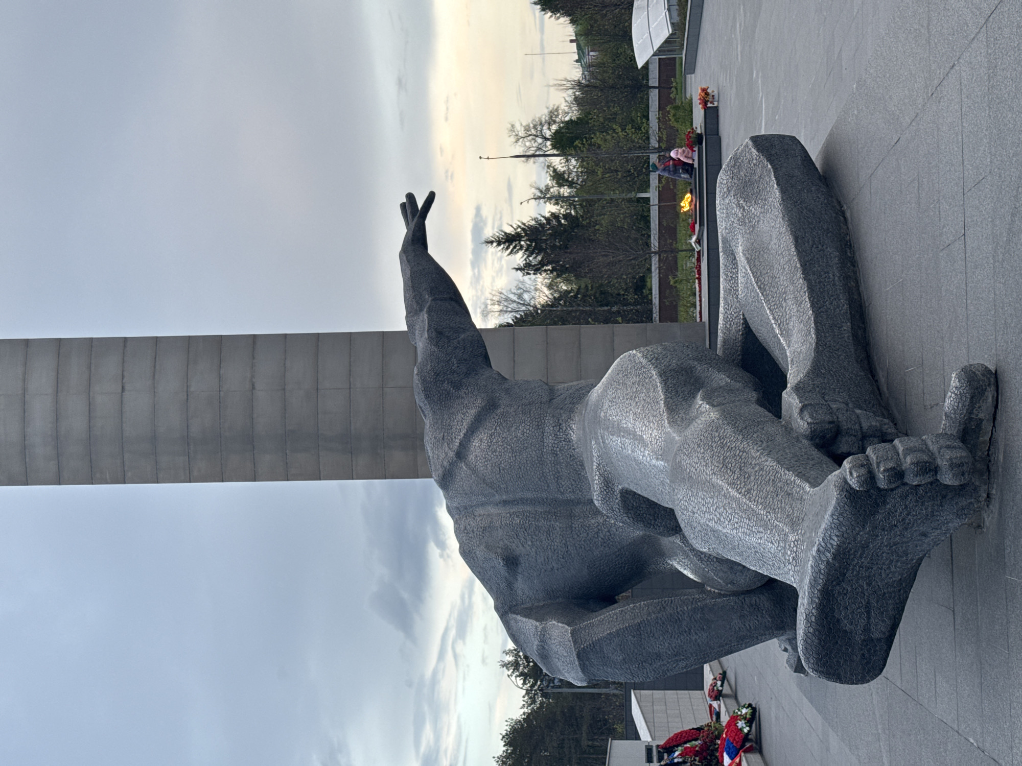 The image depicts a monument in Kazan, Russia, featuring a large, stylized stone sculpture of a kneeling figure with one arm raised. The statue appears to be positioned on a gray stone surface, possibly a plaza or memorial site. To the left, a tall, gray pillar extends upward into a cloudy sky. In the background, there are trees and greenery. Beyond that, a low fence or border encloses an area with flowers and a burning flame. Two individuals are present near the flame in the background. There is a patch of floral wreaths at the corner of the monument.