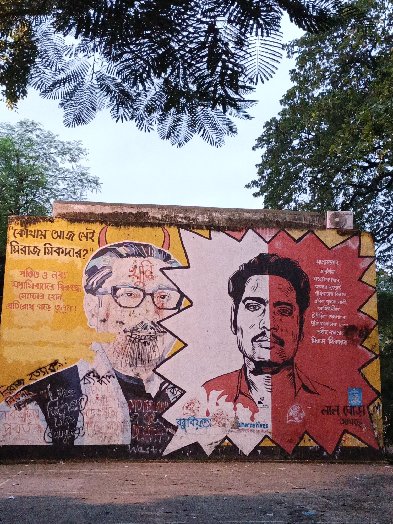The image depicts a mural painted on a yellow and red wall, likely located in Dhaka, Bangladesh, based on the Bengali text. The mural features two portraits side-by-side, separated by a jagged white line. The left portrait is of a man with glasses and a beard, depicted with devil horns, and is surrounded by Bengali text that appears critical. The right portrait is of a man with a mustache, also rendered in black and white, with the right side of his face colored red. This section of the mural also contains Bengali text and the logo for "Alternatives". Below the portraits, graffiti is visible, including English phrases like "Like father Like daughter" and "Motherland or Death", as well as the word "Fascist" and "Waster". In the background, lush green trees and a pale sky are visible, with the top of the wall showing signs of age and wear. An air conditioning unit is mounted on the wall above the right portrait. The ground in front of the mural is a paved surface, littered with small debris.