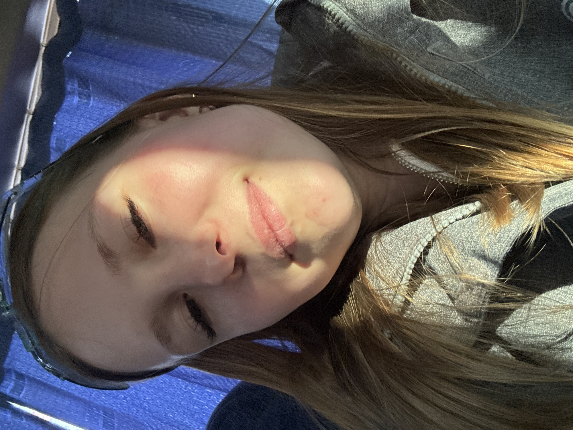 A close-up, rotated portrait depicts a young woman indoors. She has light skin, brown hair, and closed eyes. She is wearing a gray hooded jacket. Light casts shadows across her face and hair. A blue textured surface appears in the background. The location in Berezniki, Russia, is not evident from the image.