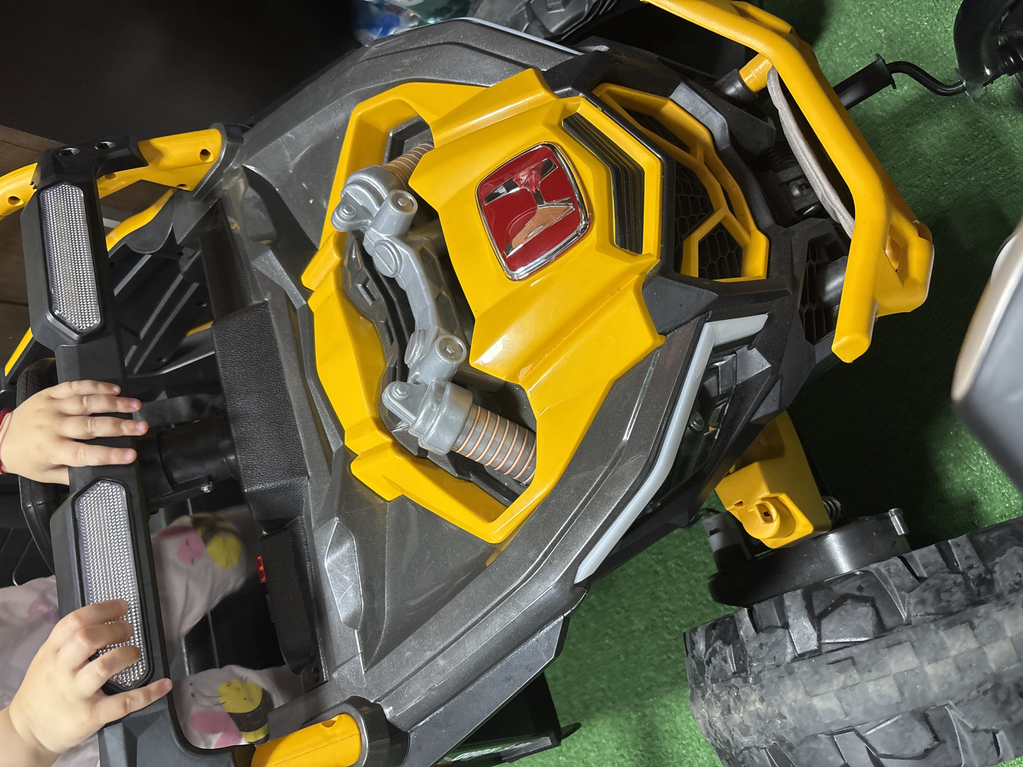The media displays a child's ride-on toy vehicle, primarily colored in bright yellow and dark grey/black, situated on a vibrant green surface, likely artificial grass or carpet. The vehicle resembles an ATV or off-road buggy, featuring rugged, dark-treaded tires and a sturdy plastic body designed with detailed mock shock absorbers and roll bar-like structures.
A child's small hands are visible on the left side of the vehicle, interacting with a textured black and clear plastic light-like component. On the front yellow panel, a prominent red square emblem, framed in chrome or silver, displays a distinct stylized silver or gold symbol, possibly a geometric "H" or a brand logo. Inside an open compartment on the vehicle, a piece of white fabric with a cartoon bee or bumblebee pattern is visible.
The scene appears to be indoors or in a covered area, with ambient lighting. The overall impression is one of a child's play item, with its robust design and the visible interaction suggesting active use. No specific text is visible other than the stylized symbol on the emblem.