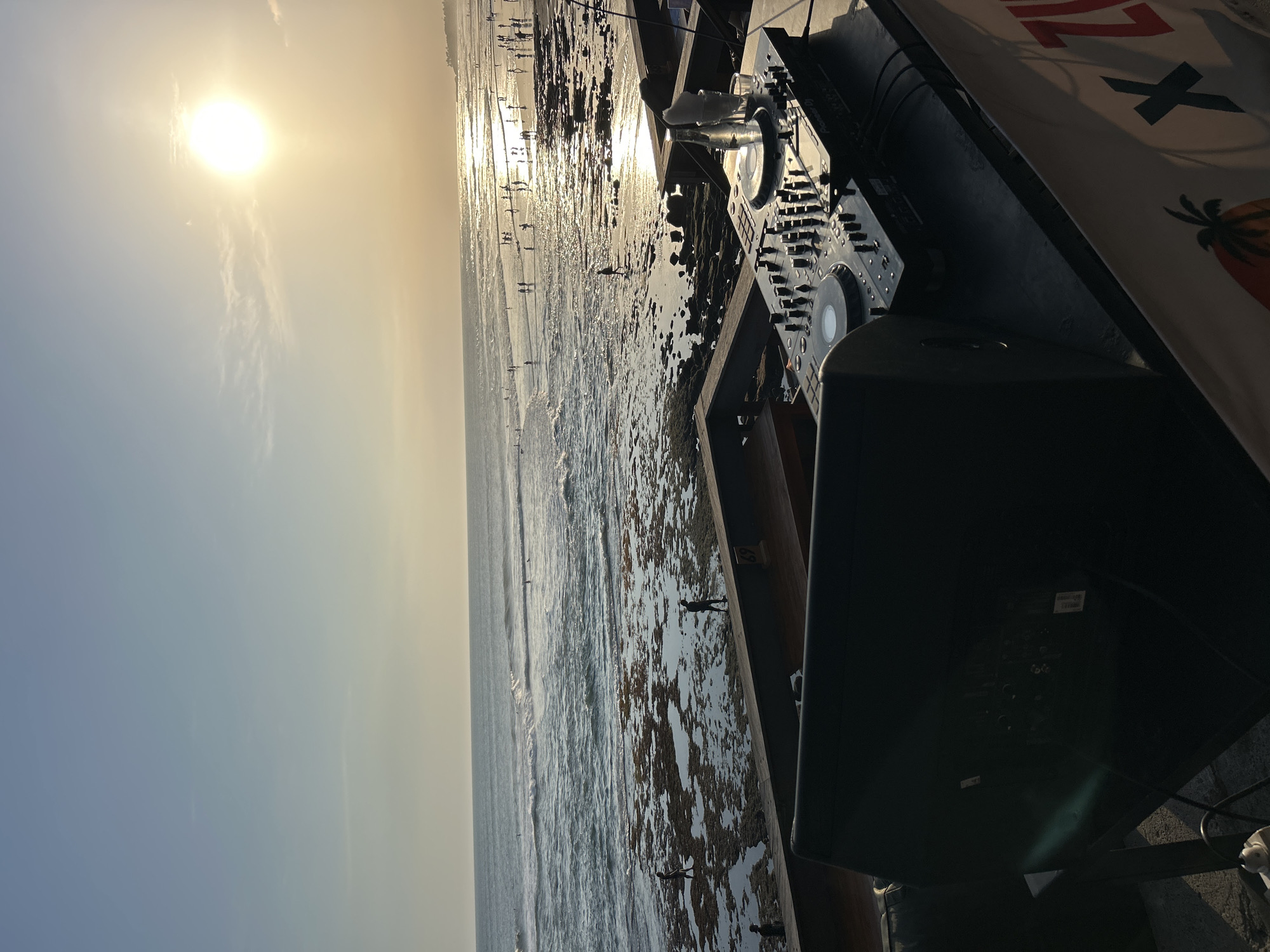 An outdoor DJ setup is positioned on a raised platform overlooking a beach at sunset. The DJ booth is equipped with a Pioneer DJ mixer and two large speakers. A bottle of water and a glass are also visible on the mixer. In the background, the ocean stretches towards the horizon, with gentle waves lapping against a rocky shore. Numerous people are scattered across the beach and in the water, some wading, others appearing to surf. The sun, a bright orb, is low in the sky, casting a warm, golden light and reflecting on the water. The scene suggests a lively evening gathering at a popular coastal spot in North Kuta, Indonesia.
