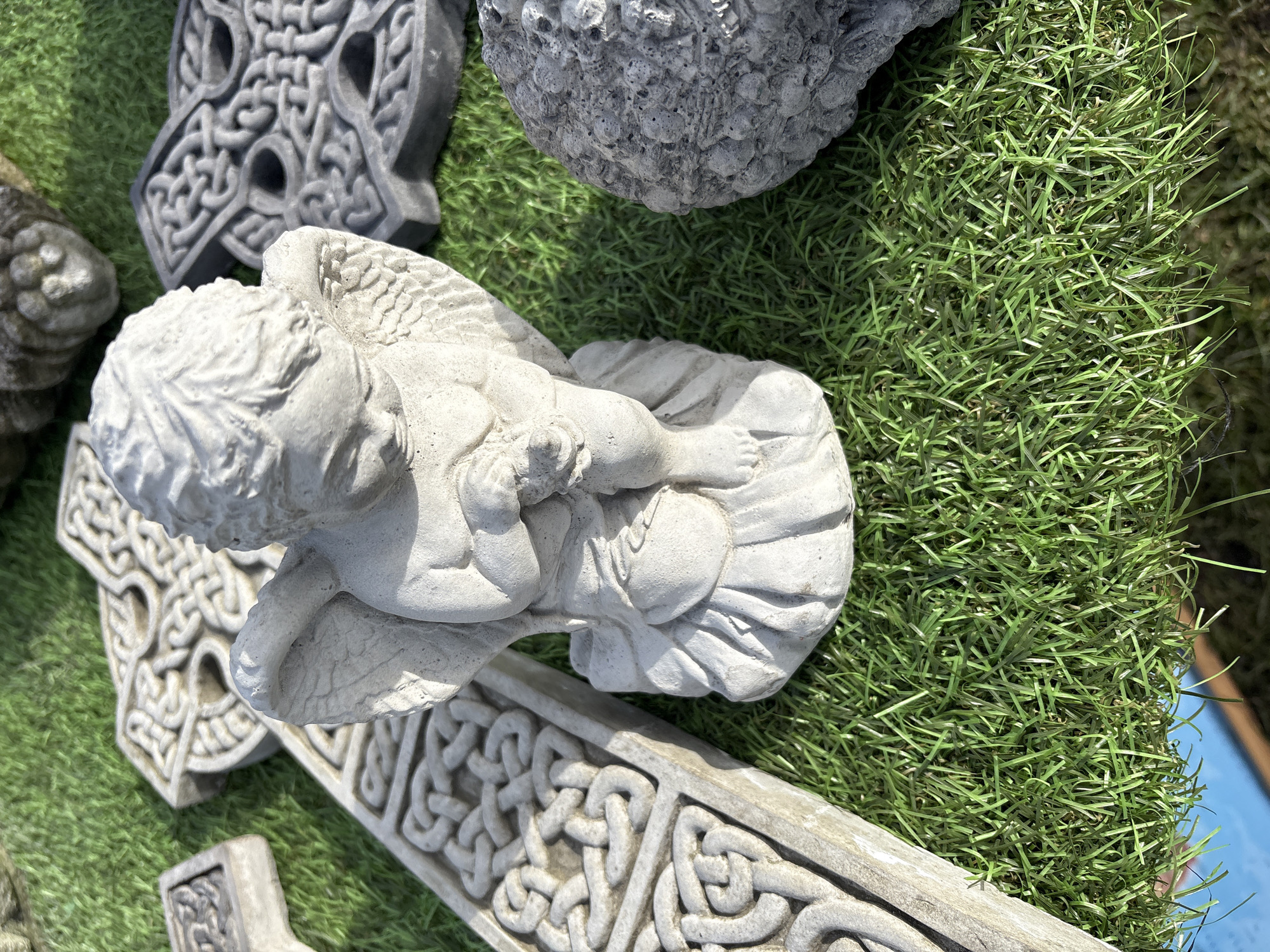 The image shows a close-up view of garden ornaments placed on artificial grass. The ornaments include a stone cherub lying on its side, a stone ball, and several Celtic crosses in various shapes and sizes. The setting appears to be outdoors, likely a garden or patio. The specific location is not evident in the image itself.