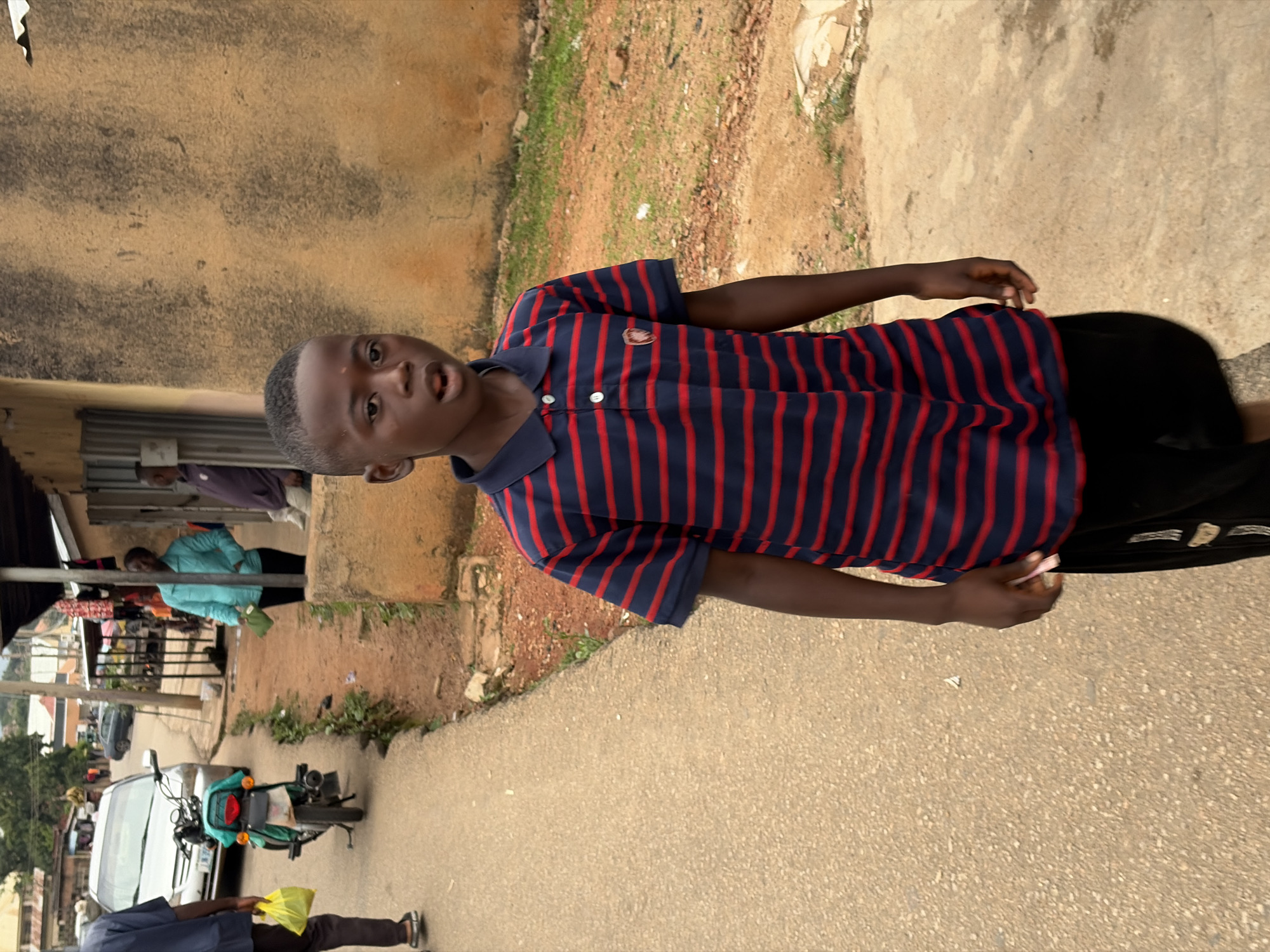 A young boy walks down a dirt road in Jos, Nigeria. He is wearing a blue and red striped polo shirt and black shorts. In the background, there are buildings, a motorcycle, and other people. The scene appears to be a typical street in a Nigerian city, with a mix of residential and commercial structures. The weather is overcast, and the road is unpaved. The boy's expression is neutral, and he is looking forward as he walks.