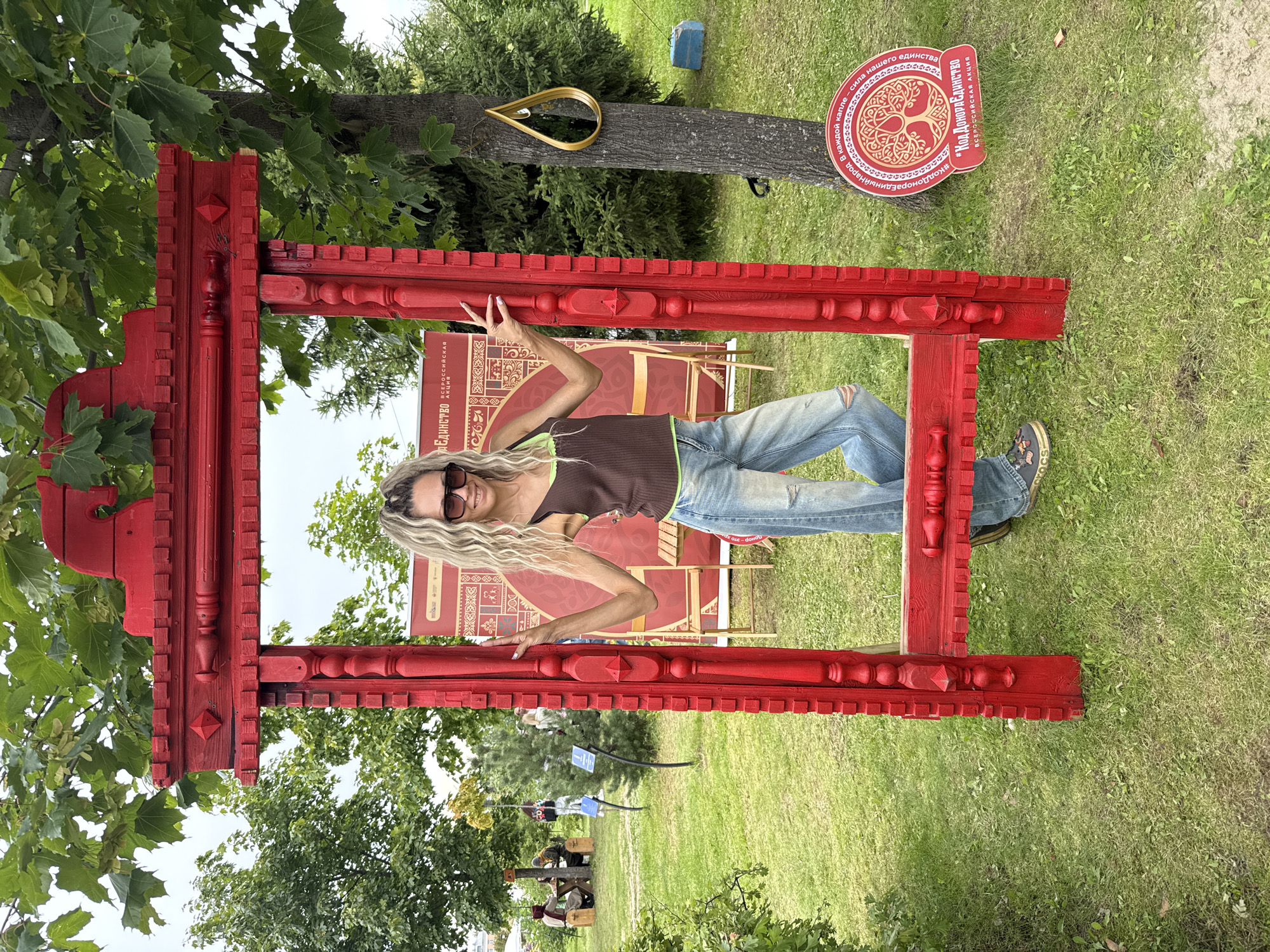 A woman with long, wavy blonde hair and sunglasses stands in the foreground, leaning against a large, ornate, red wooden frame that appears to be a photo booth. She is wearing a brown tank top with a green trim and distressed light blue jeans. Her right arm is resting on the frame, and her left arm is extended outward. Behind her, a large red banner with Cyrillic text and intricate patterns is visible. To the right of the frame, a tree trunk with a gold teardrop-shaped object hanging from it is present. Further in the background, there is a grassy area with trees, some people sitting at tables, and other structures that suggest an outdoor event or park setting. A circular red sign with the tree of life motif and Cyrillic text is on the ground to the right of the woman. The overall scene suggests a festive or cultural event taking place outdoors.
