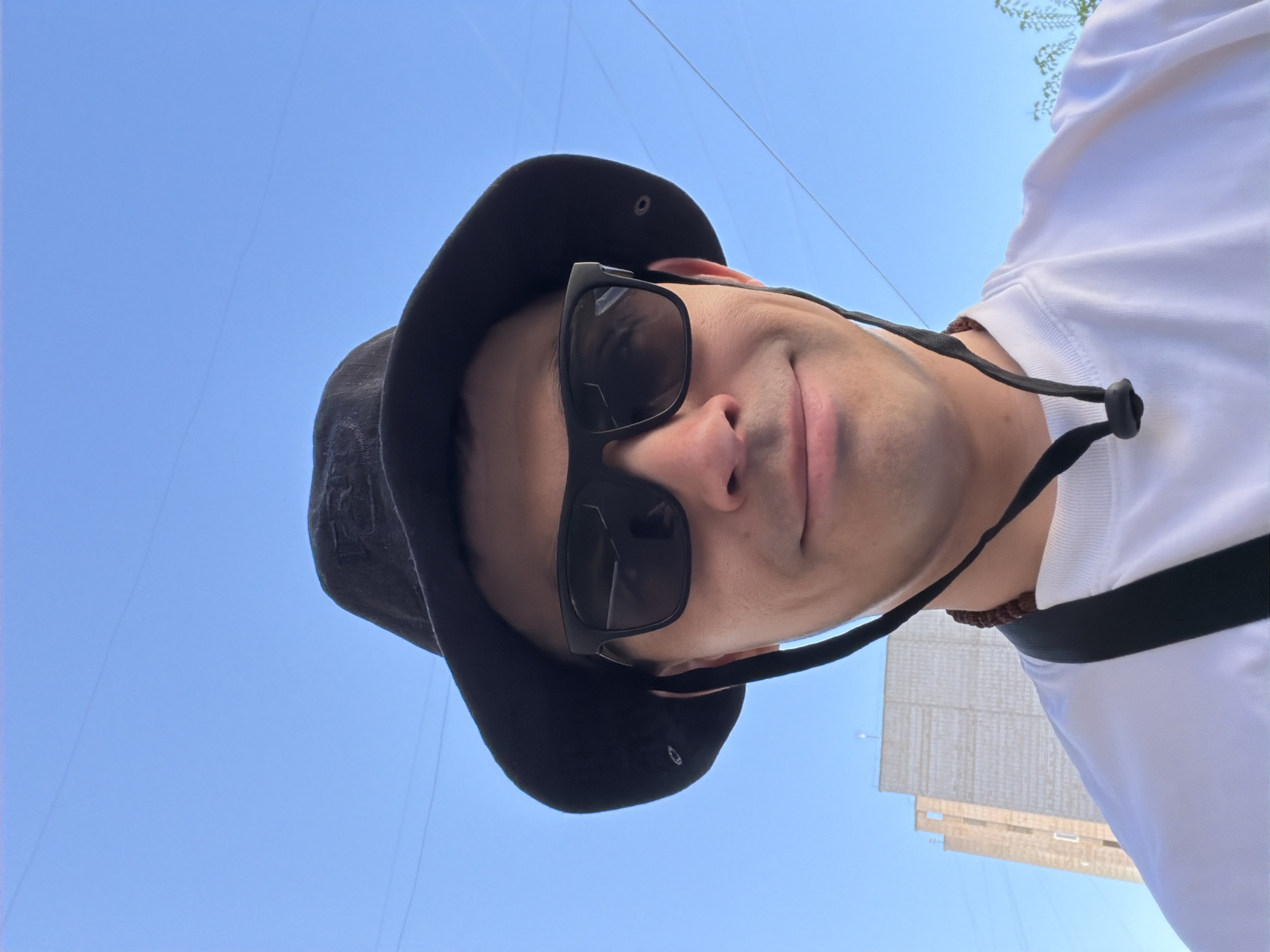 A man wearing a black bucket hat and sunglasses is taking a selfie. He is also wearing a white shirt and a black strap is visible around his neck. Behind him is a clear blue sky and the top of a light-colored building. Based on the architecture of the building and the presence of overhead wires, the location may be in Izhevsk, Russia.