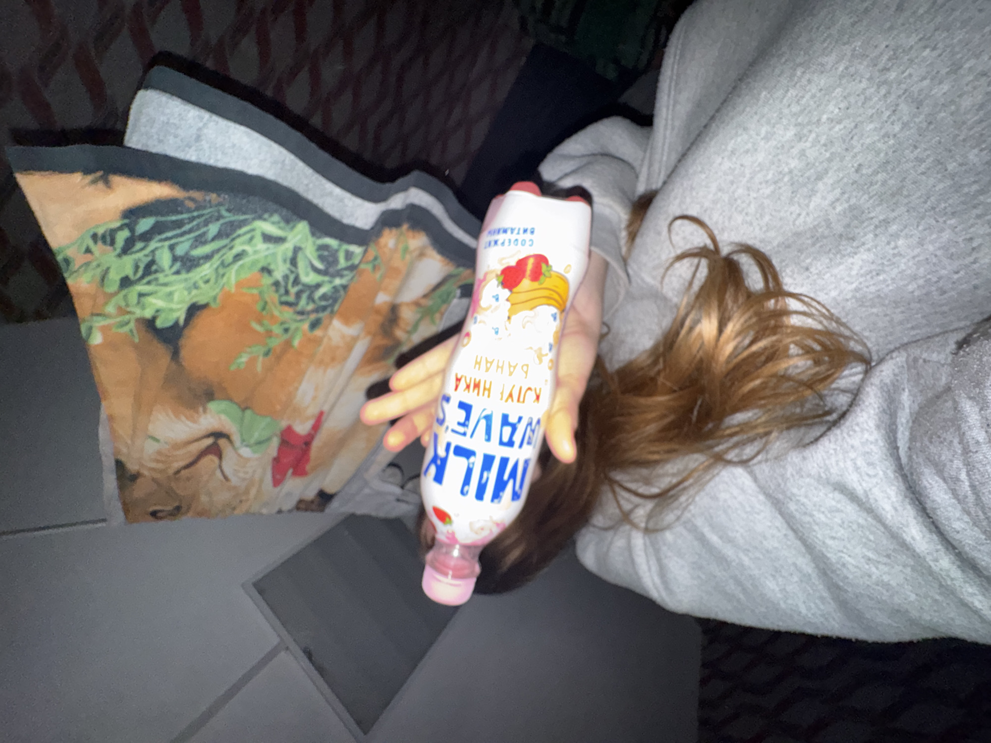 A young person is reclined with a pillow depicting a lion, holding a bottle of "Milk Waves" banana flavored milk. The bottle has text in Russian. The person is wearing a gray sweatshirt. The scene is dimly lit, seemingly indoors. Location: Perm, Russia (deduced from the Russian language on the milk bottle).