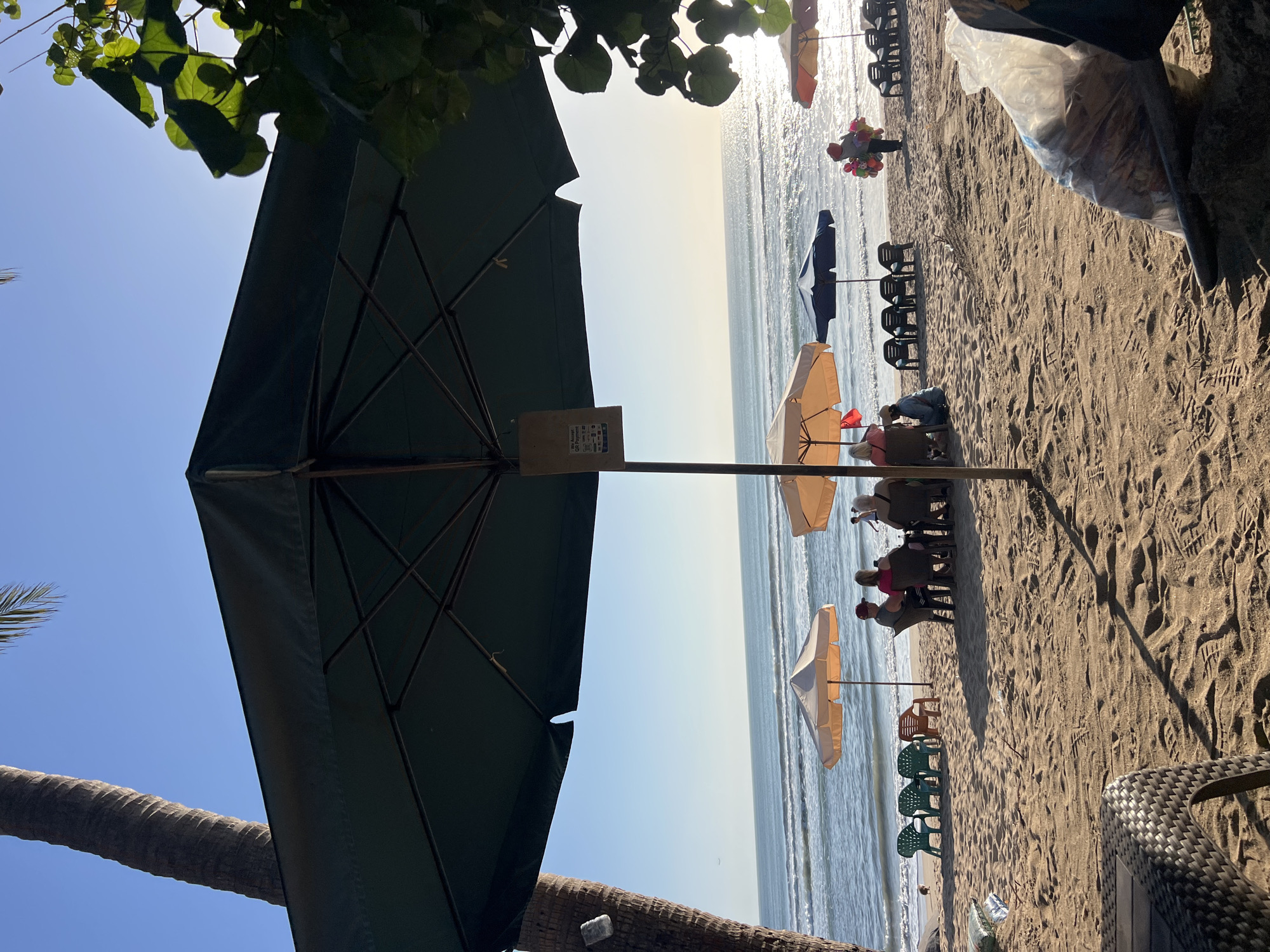 The image depicts a sun-drenched beach scene in Kuta, Indonesia. In the foreground, a large dark umbrella dominates the frame, casting a shadow over the sandy beach. To the left, a portion of a palm tree trunk is visible against a clear blue sky. Several beach umbrellas, in shades of beige, blue, and orange, are scattered across the sand, providing shade for beachgoers.
A group of people are seated around a table under one of the beige umbrellas, engaged in conversation. Further down the beach, a person is seen selling balloons, while other beach chairs are arranged in rows. The ocean, with gentle waves, stretches to the horizon, reflecting the bright sunlight. The sand is imprinted with footprints. In the far distance, a small airplane is visible in the sky. A plastic bag filled with what appears to be trash or goods lies on the sand in the lower right corner of the image. The overall atmosphere suggests a relaxed day at the beach, characteristic of Kuta's popular tourist destinations.
