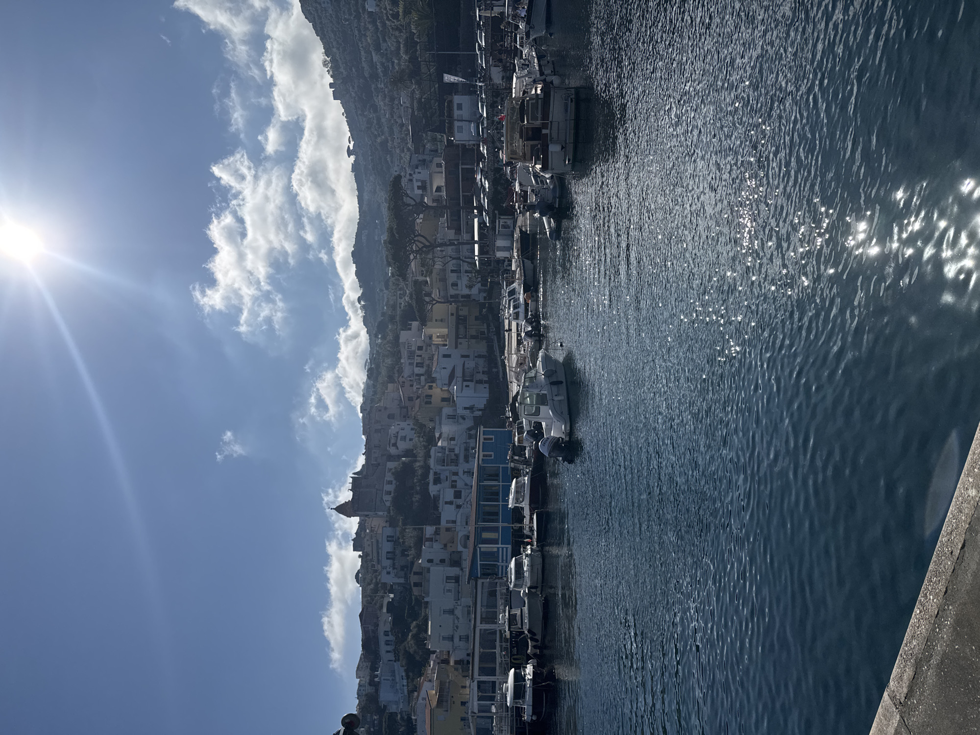 The image shows the port of Massa Lubrense, Italy. The foreground features a dark blue body of water, reflecting sunlight. A concrete structure is visible in the lower right corner. The background shows a hillside covered in buildings and vegetation. A variety of boats are docked along the shoreline. The sky is partially cloudy with the sun shining brightly in the upper left corner.