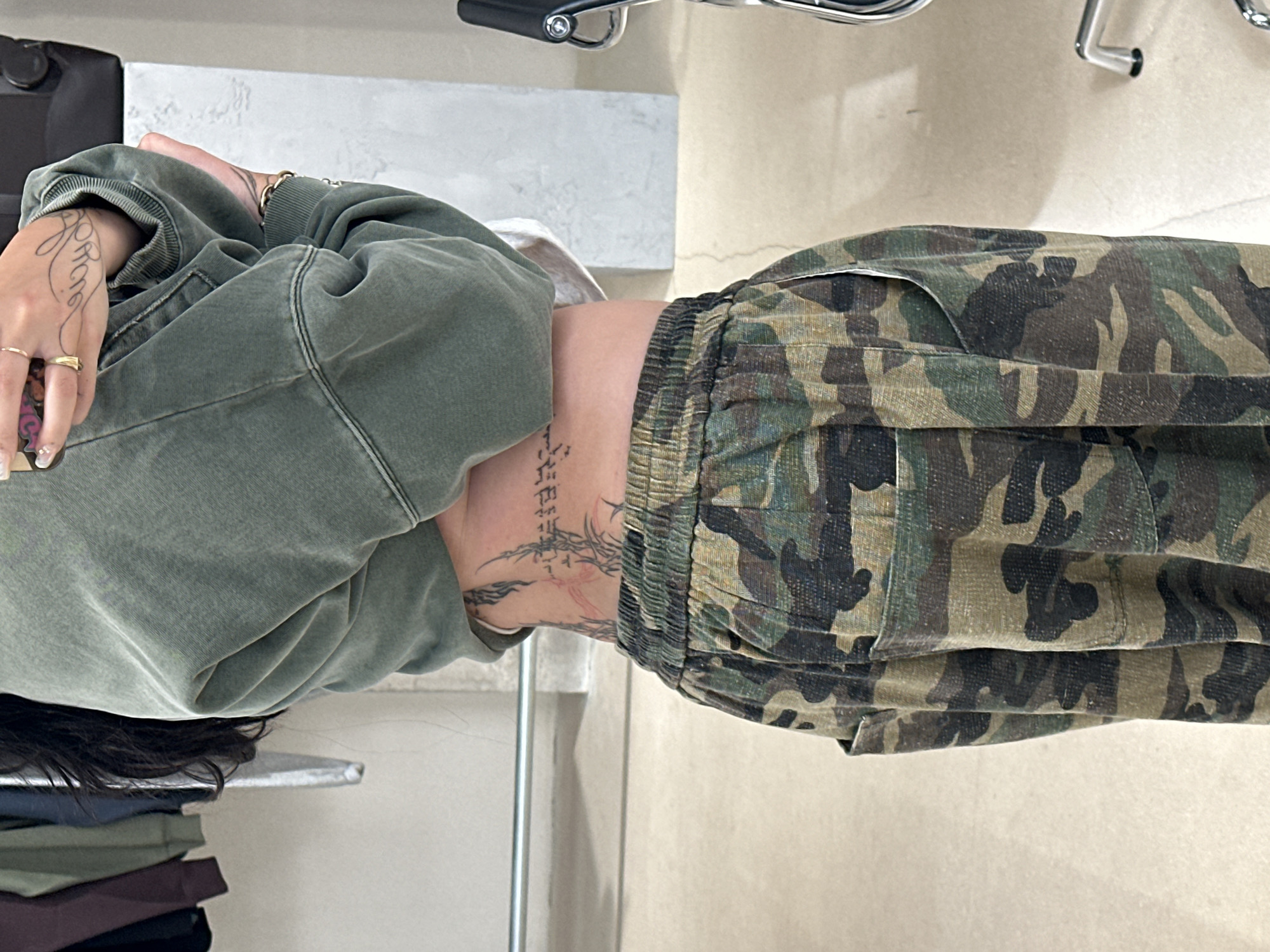 A person wearing a green sweatshirt and camouflage pants is shown in a medium shot. The person is sitting in an indoor location with a concrete-like shelf to the left and a cream colored wall to the right. There are additional garments folded on the shelf. A tattoo is visible on the person's wrist, and another runs vertically down the torso. A chair is present in the upper portion of the photo. The location is indeterminate.