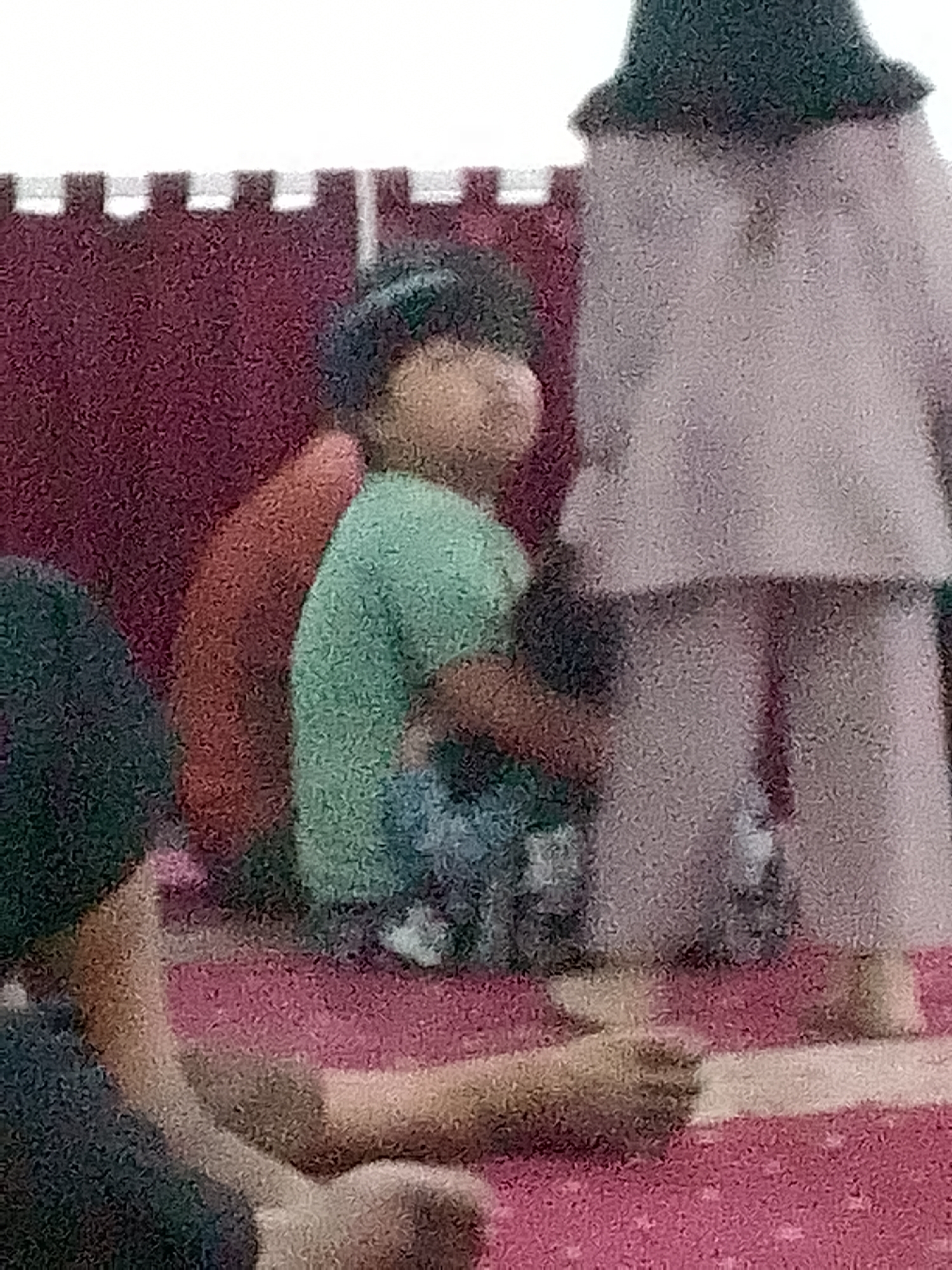 The image depicts a dimly lit interior scene, likely a room with red carpeted floor. In the foreground, a person's arm is visible, resting on the carpet. Further back, a young child wearing a green shirt and dark cap is seated, leaning against a dark red object. Behind the child, a person in a light-colored outfit, possibly a dress or robe, stands facing away from the camera. The background appears to be a patterned wall or partition. The overall image quality is grainy, suggesting it was taken in low light or with a low-resolution camera. The context of Gunungmasigit, Indonesia is not visually discernible from the image itself.