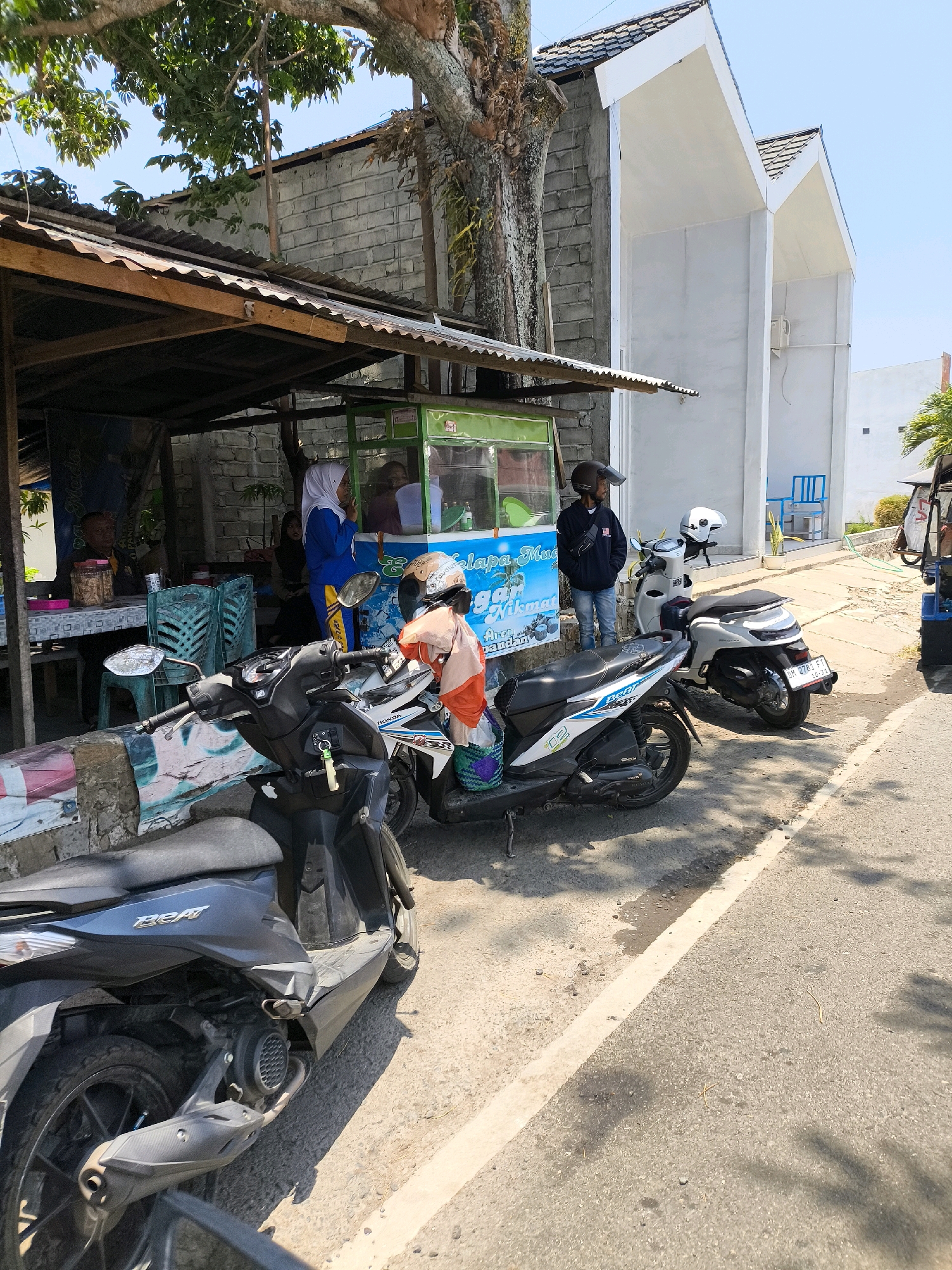 This image depicts a street scene in Kwandang, Indonesia, on a bright, sunny day. In the foreground, two scooters are parked on a paved road with a white line marking the lane. The scooter on the left is dark gray and a portion of its rear is visible. The scooter on the right is white with blue and orange accents, and a person wearing an orange shirt and a helmet is seated on it, holding a bag.
Behind the scooters, a small, open-air food stall is set up under a corrugated metal roof supported by wooden beams. A brick wall forms the backdrop of the stall. Inside the stall, a woman wearing a blue hijab and a blue and yellow tracksuit is standing and appears to be interacting with someone behind a green display case. Another person is partially visible in the background, seated at a table. Several plastic chairs are arranged around tables.
To the right of the food stall, a modern, two-story white building with a distinctive peaked roof design is visible. A blue plastic chair is placed outside the building on a paved area. The environment suggests a typical urban or semi-urban setting in Indonesia, likely a commercial or residential area. The overall impression is one of everyday life, with people engaged in activities around a local eatery and the street.