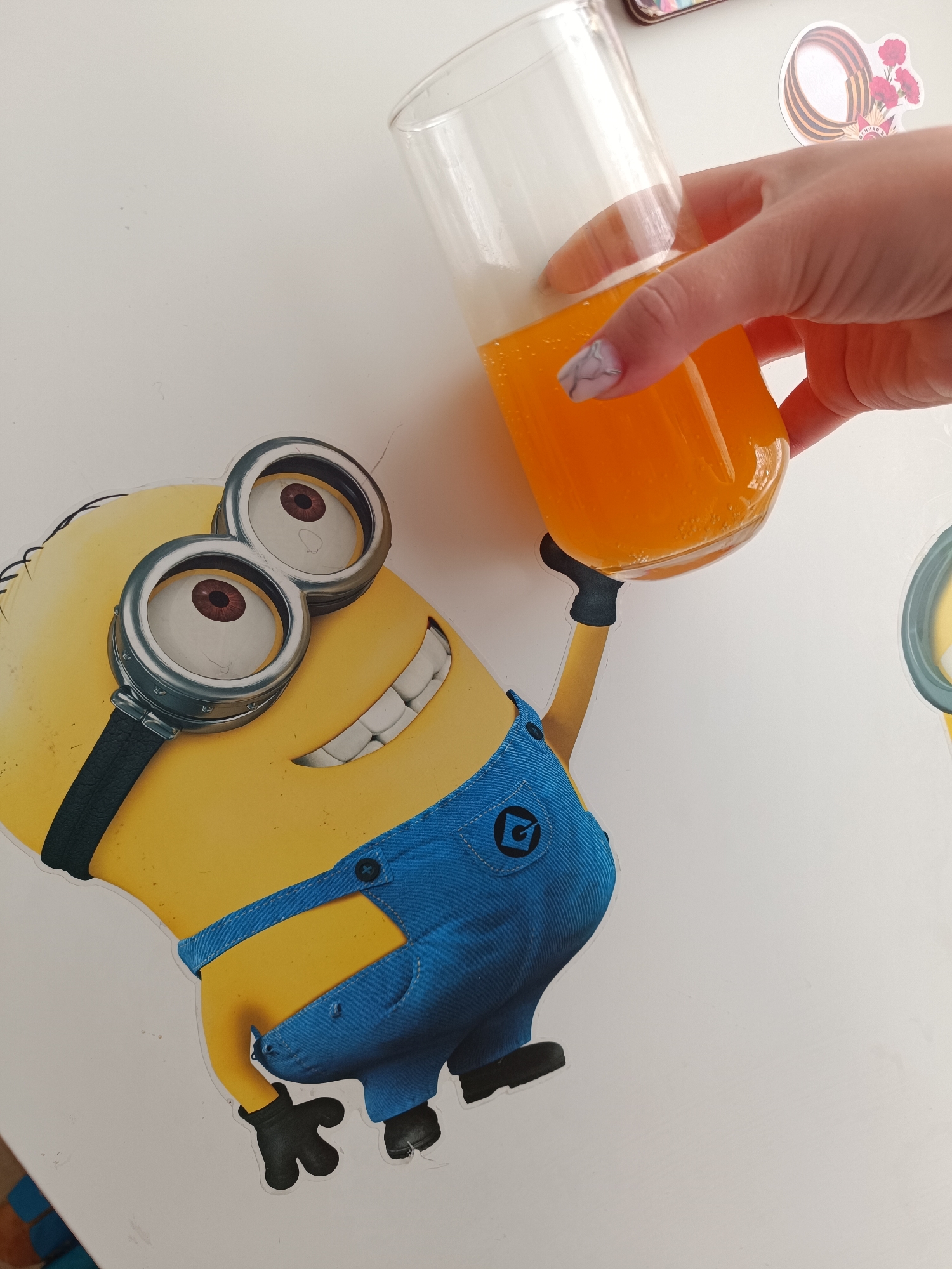 A photograph depicts a Minion cartoon character holding a glass of orange liquid. The scene is brightly lit, with the Minion placed on a white surface. A hand holds the glass, appearing to offer a toast. The setting lacks specific contextual indicators tying it directly to Tobolsk, Russia.