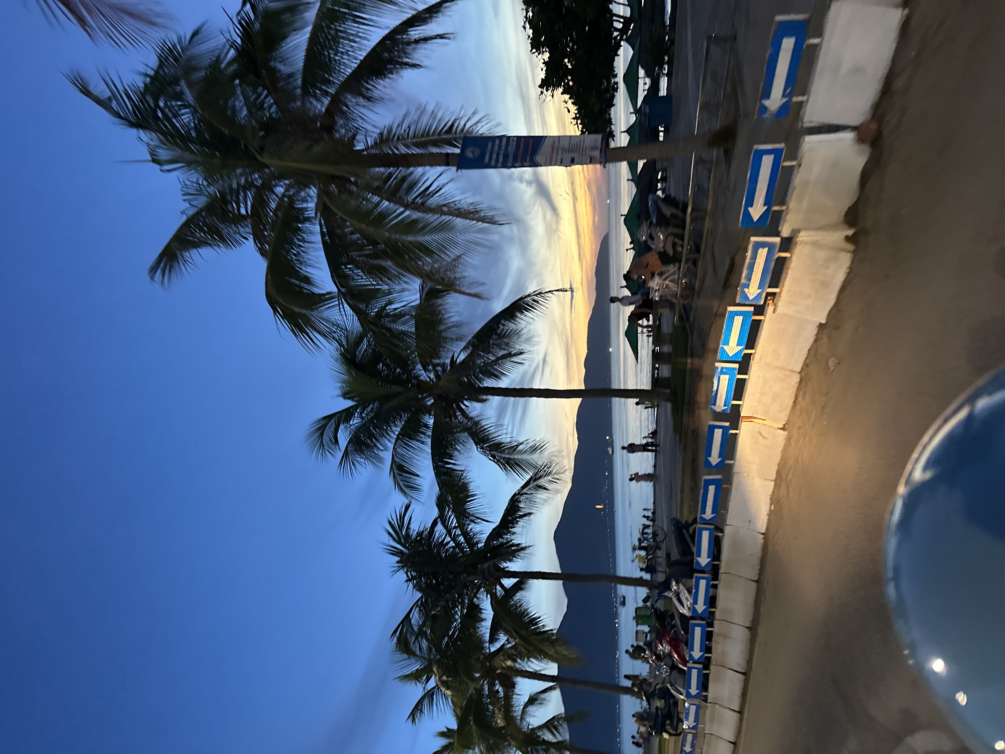 The image captures a twilight scene on a beach promenade in Da Nang, Vietnam. The sky is a gradient of deep blue to orange and yellow hues, indicating the sunset. Several palm trees line the promenade, their fronds silhouetted against the sky.
In the foreground, a paved road is visible, with a blue and white barrier along its edge, marked with numerous blue signs displaying white arrows pointing to the left. Several motorcycles are parked behind the barrier.
Further back, the beach is populated with various people. Some are strolling along the shore, while others are seated or standing near beach umbrellas, suggesting a recreational area. The sea stretches out to the horizon, where a range of mountains can be seen. The lights of a city are faintly visible on the distant coastline. The overall atmosphere is tranquil, with the fading light creating a serene mood.