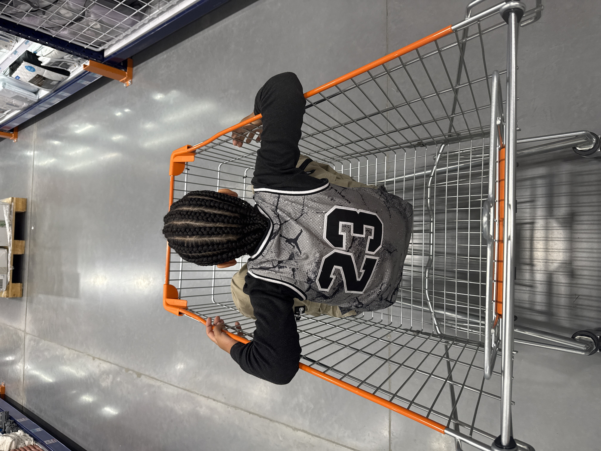 The image shows a child sitting in a shopping cart in a large store with a concrete floor and shelves lining the walls. The child is facing away from the camera and is wearing a grey jersey with the number "23" on it. The shopping cart is made of metal with orange accents on the handles. The shelves on the right side of the image appear to be stocked with various products. The overall environment suggests a retail setting, possibly a department store or a supermarket. The location is identified as Mosquera, Colombia.