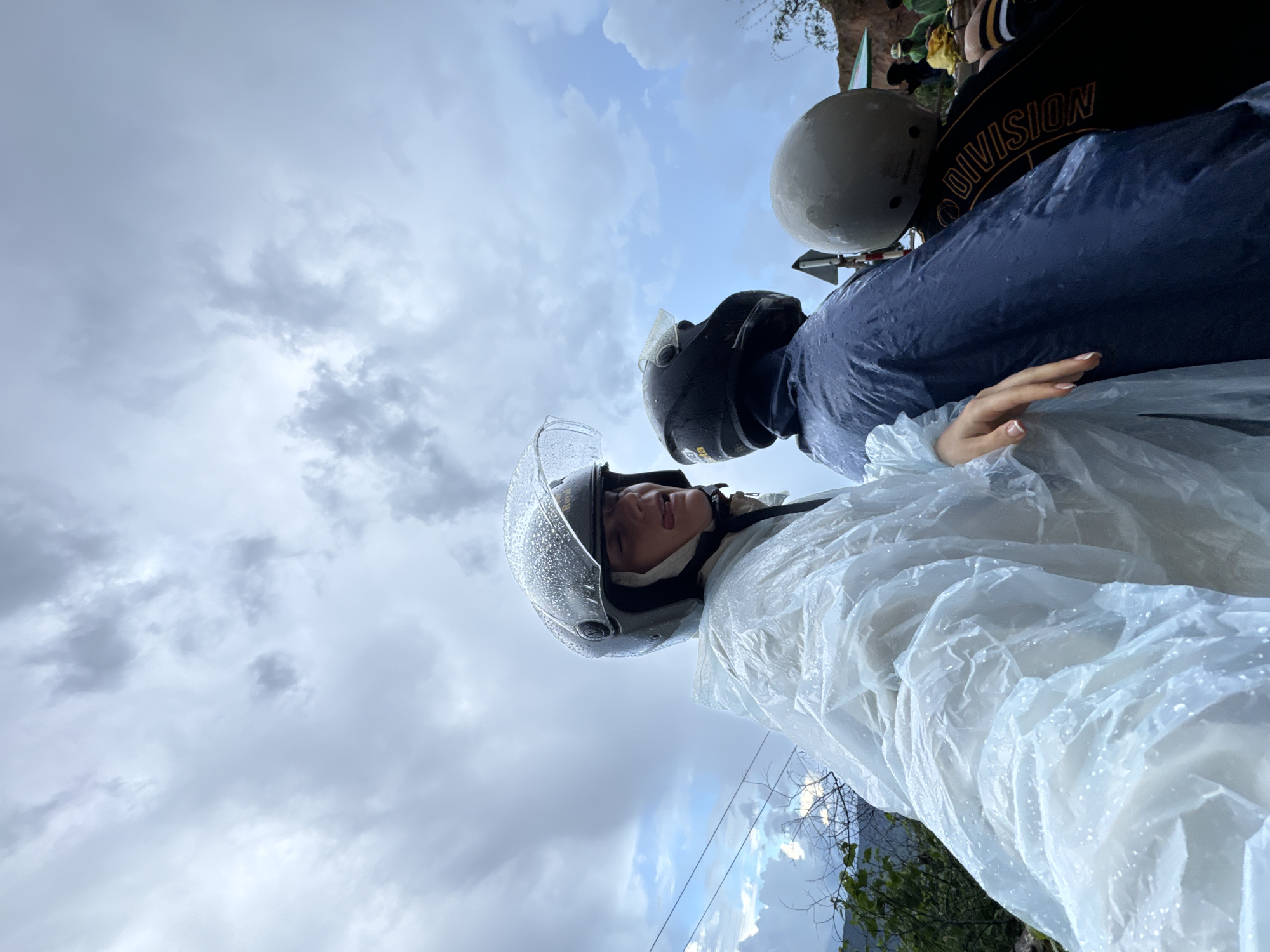 The image shows three people on a motorcycle in Nao Sẻ, Vietnam, wearing raincoats and helmets as it is raining. The person in the foreground is sticking their tongue out playfully, with water droplets on their clear helmet visor. The person in the middle is wearing a dark blue raincoat and a black helmet. The third person, mostly obscured, is wearing a light grey helmet. The background shows a cloudy sky and some greenery. The overall atmosphere is casual and adventurous, despite the rainy weather.