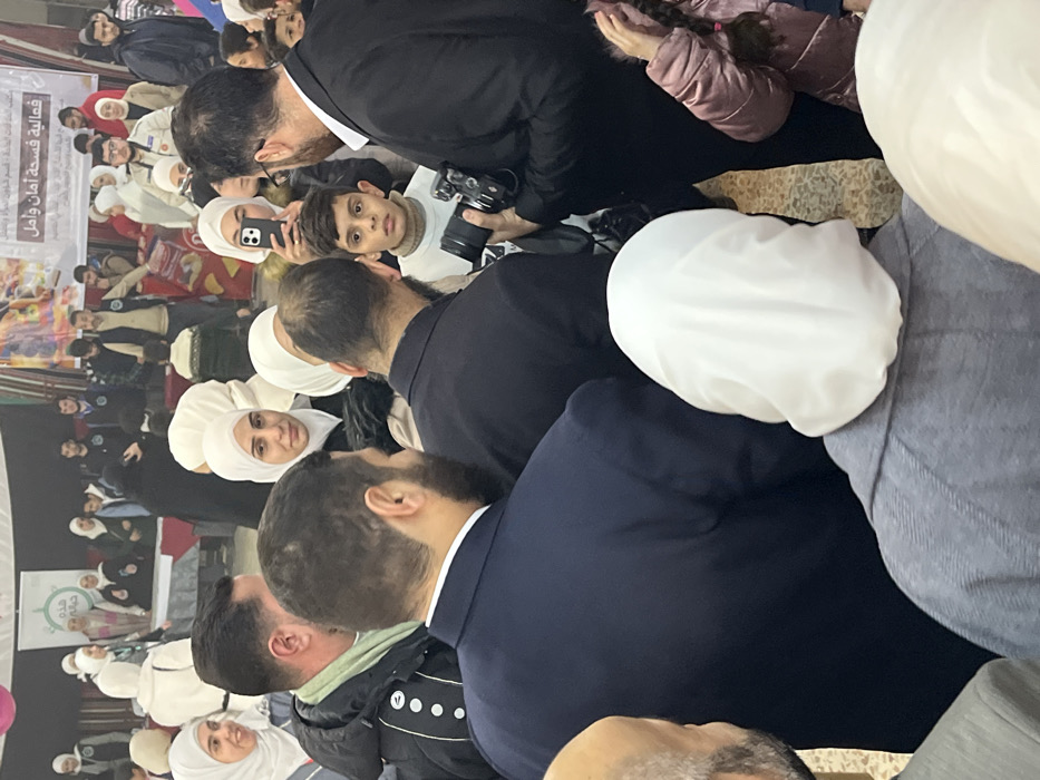 This image captures a crowded indoor event in Kafr Batna, Syria. Approximately 30-40 individuals are visible, predominantly adults. In the foreground, the backs of three individuals are prominent: a man in a dark navy suit on the left, another man in a dark suit to his right, and a woman in a grey coat with a white hijab on the far right.
In the mid-ground, a young boy with short dark hair, wearing a light beige turtleneck sweater, looks intently towards the left. To his right, a man in a dark suit holds a black DSLR camera with a substantial lens, pointing it towards the boy or the central activity. Another woman, wearing a white hijab, is positioned behind the boy, holding a smartphone with a light-colored case up as if recording. Numerous other people, including women wearing hijabs and men with beards, are present throughout the scene, observing the activity.
The background features a large white banner stretched across a wall, displaying Arabic text that translates to "An event of Safety and Hope." A smaller banner with green text is partially visible on the far left. Beneath the banners, several individuals stand behind tables covered with red and white tablecloths, indicating display or information booths. The flooring appears to be a light-colored tiled surface. The overall environment suggests a community gathering or organized public event.