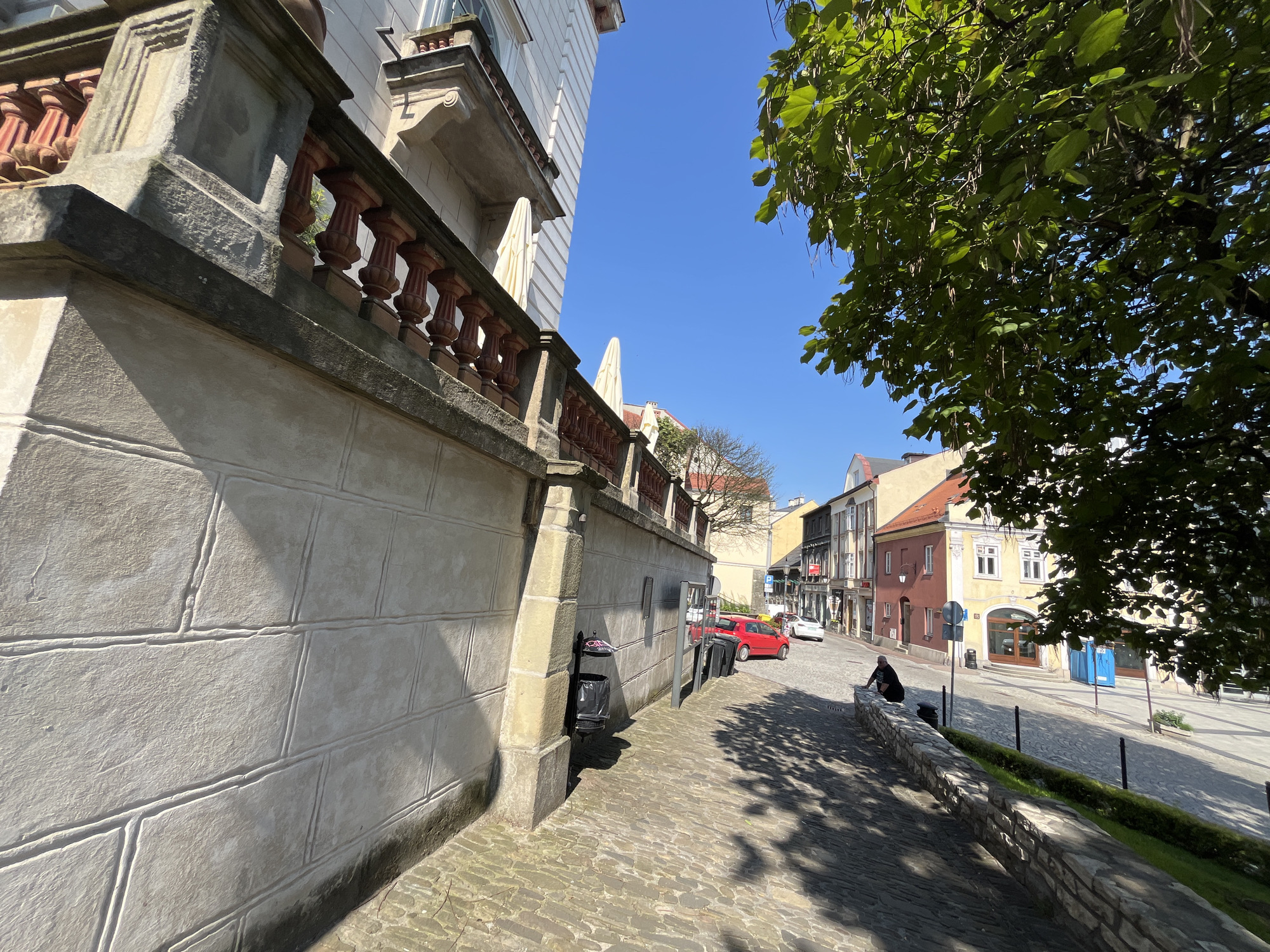 The image depicts a street scene in a city, possibly in Europe, given the architecture and cobblestone street. In the foreground, a stone wall with a balustrade runs along the left side, casting shadows. A cobblestone path leads away from the viewer, lined with a stone wall and greenery. A trash can is visible near the stone wall. Further down the street, several buildings with colorful facades line the road. A red car is parked on the right side of the street, with a white car behind it. A person is sitting on the stone wall, facing away from the camera, on the right side of the street. A tree with green leaves hangs over the scene on the right. The sky is clear and blue.