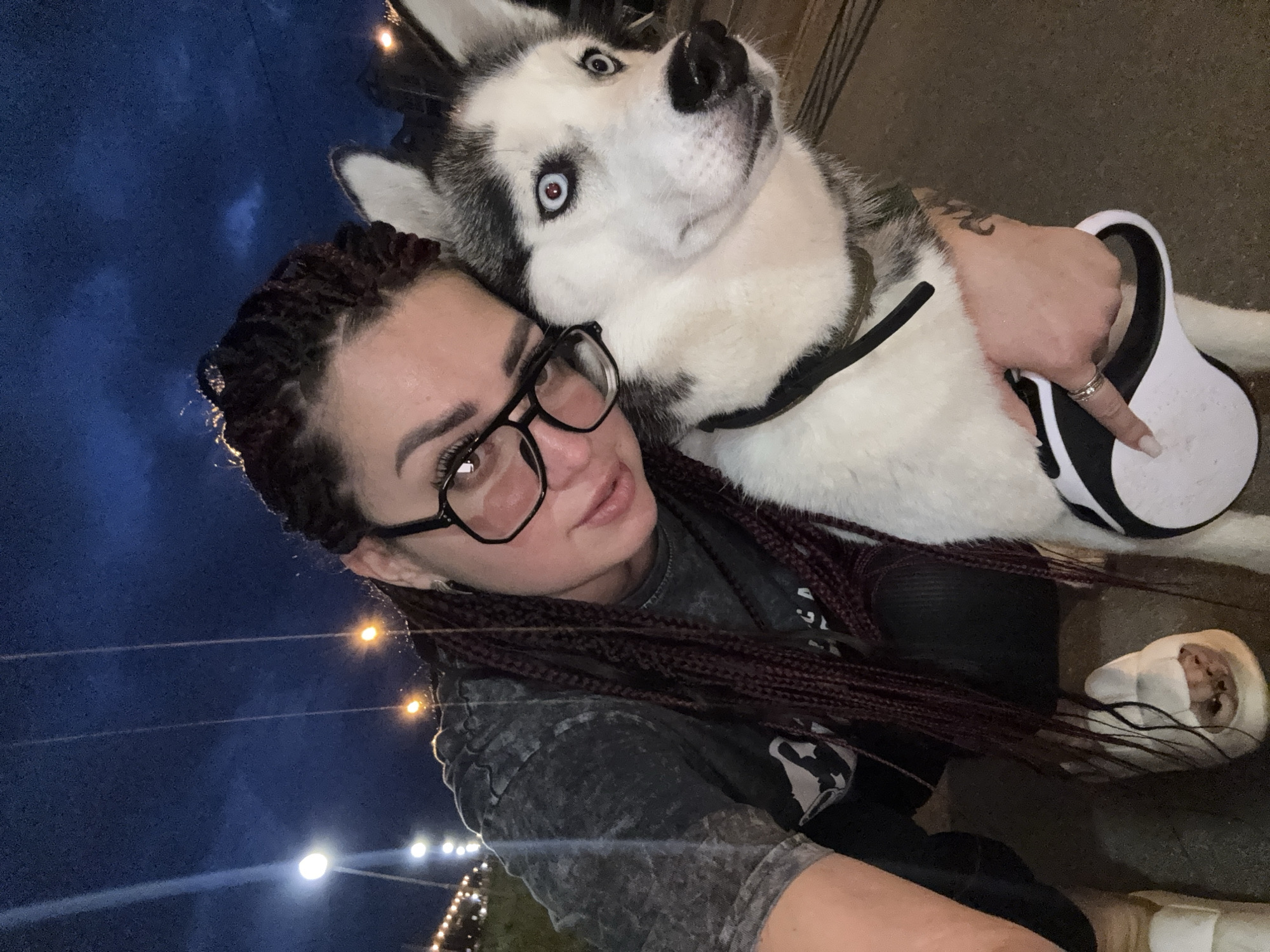A woman with braided, dark red hair and glasses is embracing a husky dog. The dog has striking blue eyes and is looking off to the side. The woman is wearing a dark grey t-shirt. They are outdoors at night, with streetlights visible in the background, suggesting an urban environment. The ground appears to be paved. The woman is holding a retractable dog leash. The overall scene suggests a casual moment between the woman and her dog.