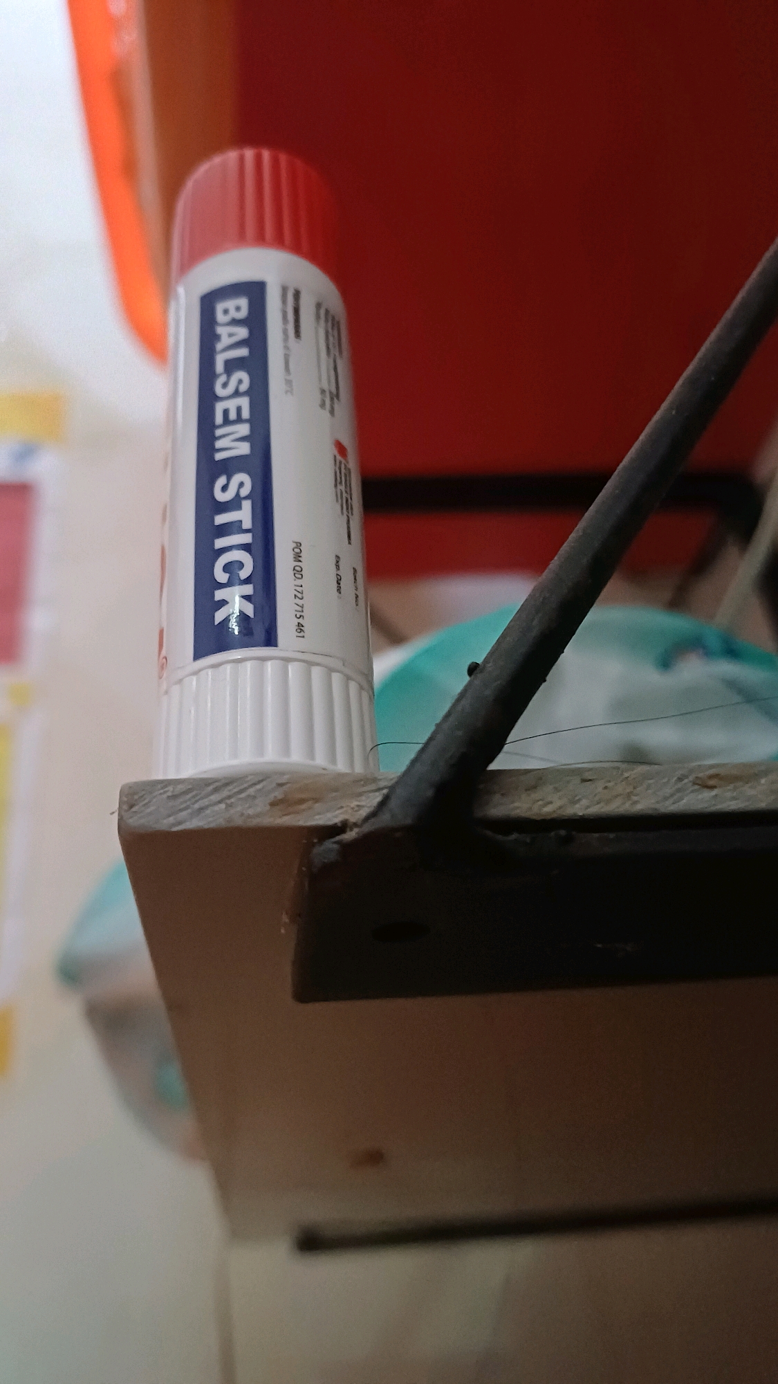 A close-up, angled shot focuses on a white tube labeled "BALSEM STICK" with a red cap. The tube is positioned on the edge of a wooden shelf, which is dark brown and has a rough texture. A thin, dark metal rod protrudes from the shelf to the right, angled upwards and away from the camera. In the background, out of focus, are hints of red, orange, and a teal object. The scene appears to be indoors, possibly in a home or shop, with no immediate indication of its specific location within Wonokerso, Indonesia, or any people present.