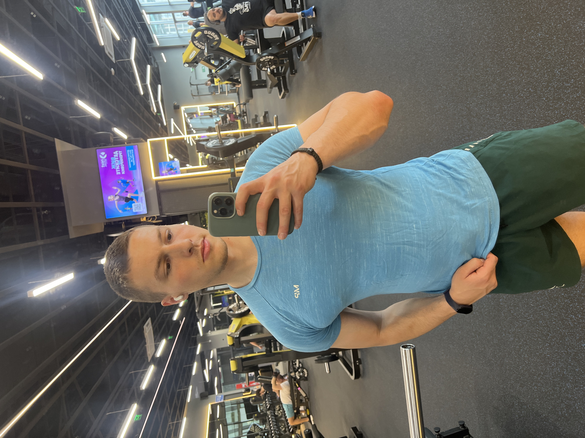 A man is taking a selfie in a gym. He is wearing a light blue t-shirt and dark green shorts. He has a fitness tracker on his left wrist and a bracelet on his right. He is holding an iPhone to take the picture. The gym has various weight machines and equipment, with other people working out in the background. A large screen is showing a program. The image was taken at a gym in Burgas, Bulgaria.