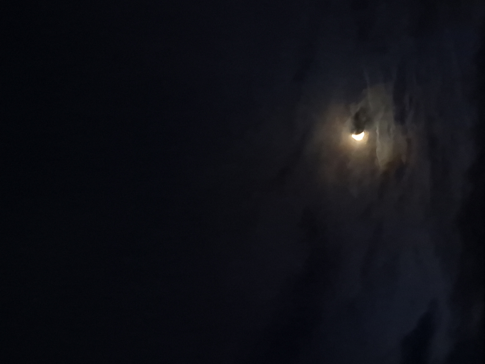 A thin crescent moon is visible in the night sky, partially obscured by wispy clouds. The moon is illuminated from the left, casting a soft glow on the surrounding clouds. The overall scene is dark and moody, with a sense of mystery. The location is the city of Sooblase, Estonia.