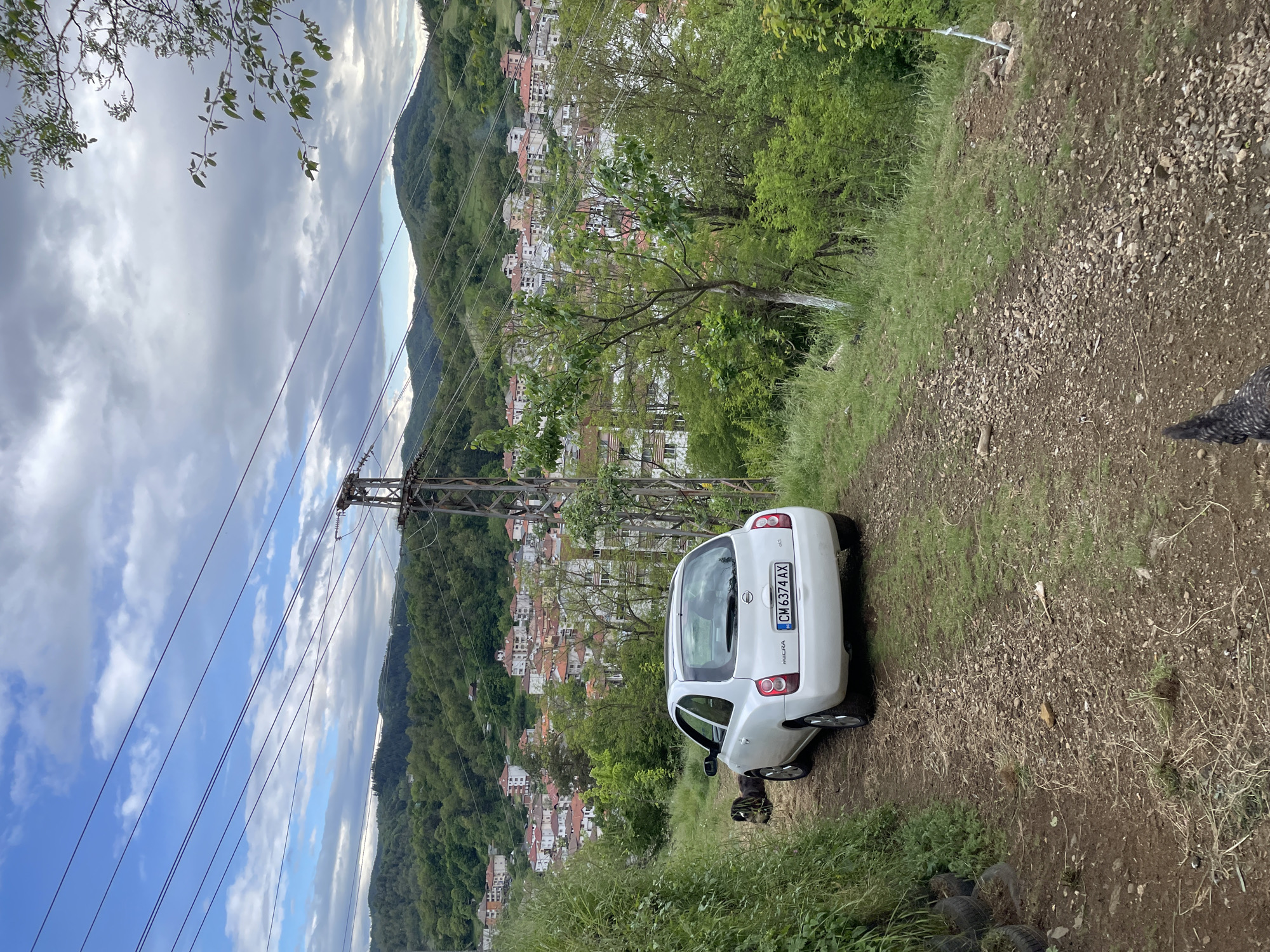 The image depicts a white Nissan Micra car parked on a dirt road overlooking the city of Zlatograd, Bulgaria. The car's license plate reads "CM 6374 AX". The city is nestled in a valley, surrounded by forested hills. Overhead power lines and a utility pole are visible in the foreground. The sky is overcast. Tires are piled on the left. Part of the tail of a chicken is visible on the right.