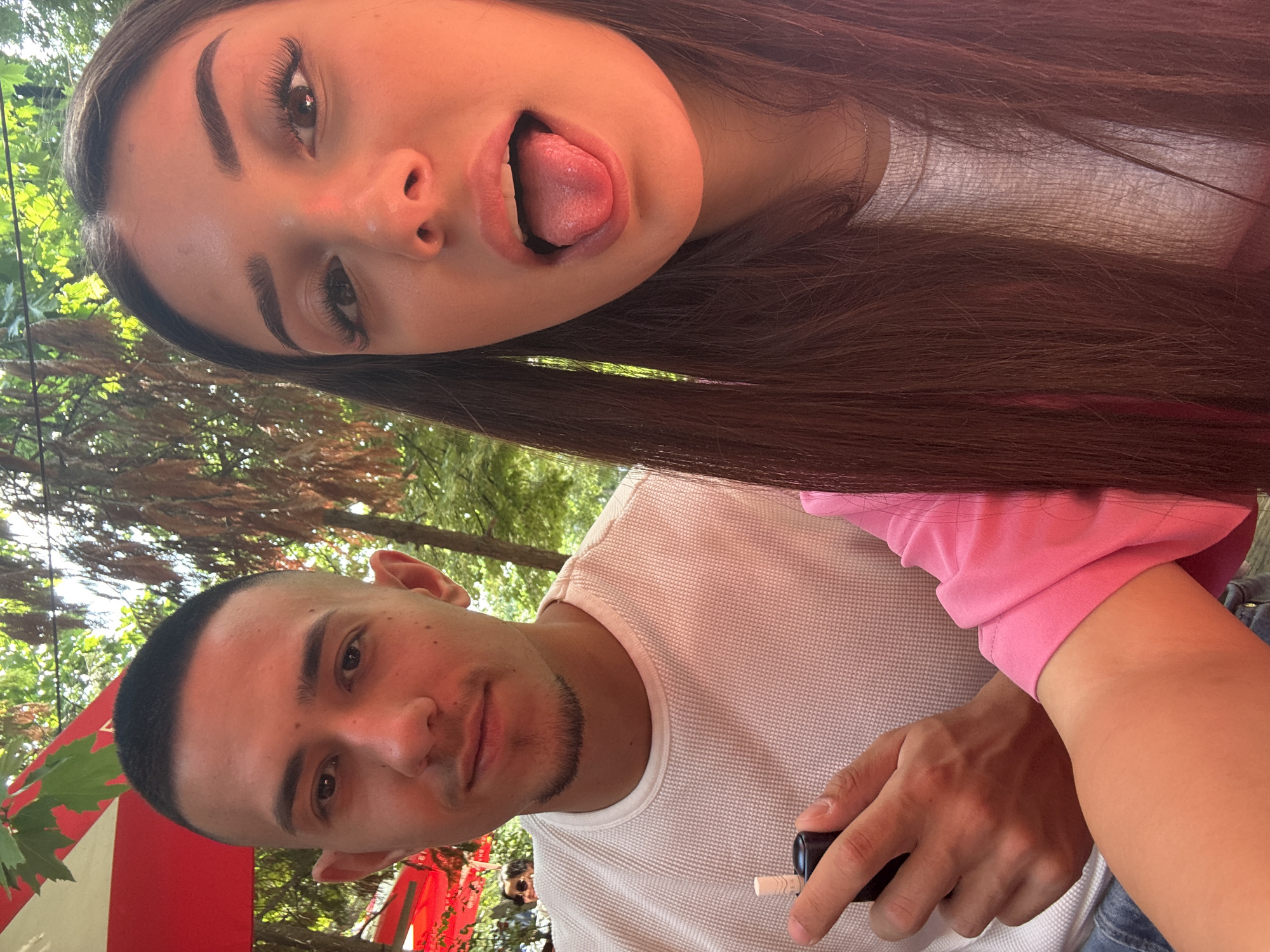 An informal close-up selfie features two young individuals in an outdoor setting, likely a cafe or restaurant in Plovdiv, Bulgaria. The image is rotated 90 degrees clockwise, with the subjects appearing tilted to the left.
In the upper portion of the frame, a young woman with long, dark hair, prominent eyebrows, and long eyelashes makes a playful expression, sticking her tongue out. She is wearing a light-colored top under a bright pink long-sleeved garment, with only parts of it visible.
Below her, a young man with short dark hair and a neatly trimmed goatee looks directly at the camera with a neutral expression. He is dressed in a white waffle-knit long-sleeved shirt. In his right hand, he holds a small black object with a white tip, which appears to be a vaping device or an inhaler.
The background shows green foliage from trees and hints of red canopies or umbrellas, characteristic of an outdoor dining area. The bright, natural lighting indicates it is daytime. No text is visible in the image.