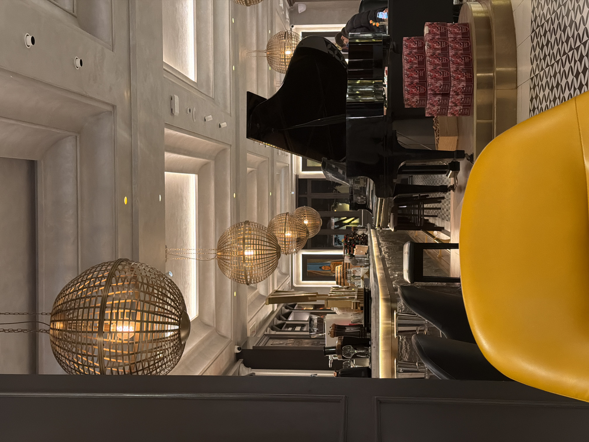 The image shows the interior of a building in Qinhuai District, China. The focal point is a grand piano situated near a bar area. Several spherical light fixtures hang from the ceiling. There are bar stools positioned along the bar, with a prominent yellow chair in the foreground. Stacks of boxes are visible near the piano. The architectural style includes recessed lighting and paneled walls. People are present in the image, though their actions are not distinctly visible. The environment is dimly lit.