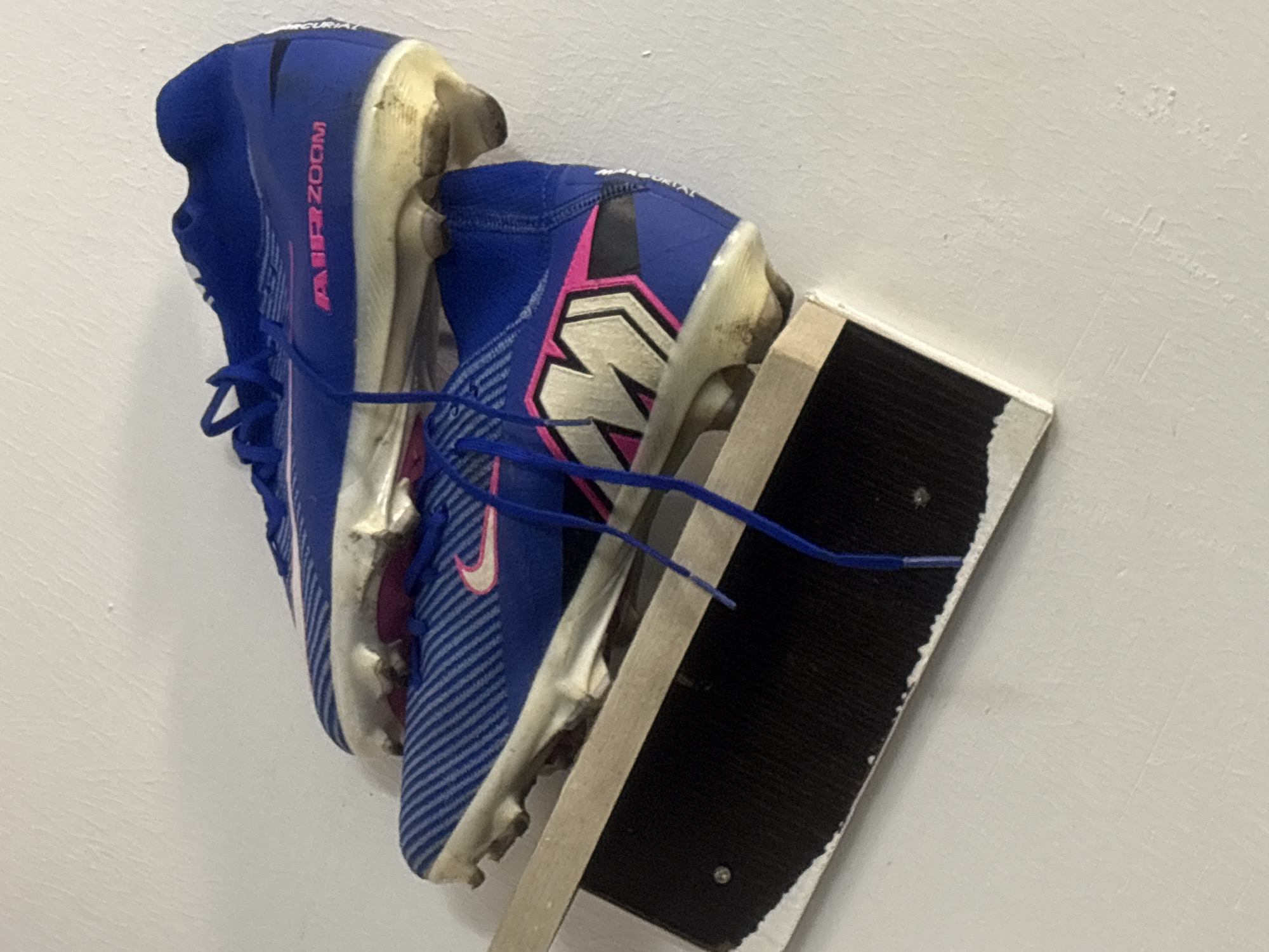 Two blue and pink soccer cleats are mounted on a white wall, supported by a wooden frame with a black backing. The cleats are worn and dirty, suggesting use. "AIR ZOOM" is printed on the side of one cleat. The frame appears to be a makeshift display. The scene is indoors, likely a residence or storage area. There is no indication of the location being Gura Top, Nigeria.