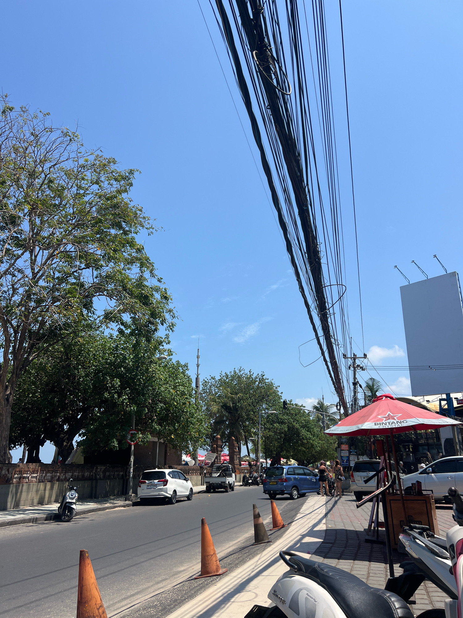 The image depicts a street scene in Kuta, Indonesia, on a clear, sunny day. The foreground features orange traffic cones on the side of the road and a parked white scooter. The street itself is paved and appears to be a busy thoroughfare, with several vehicles visible, including a white SUV, a blue minivan, and a white pickup truck. A white car is parked on the side of the street.
On the sidewalk, a red umbrella with "Bintang Crystal" branding provides shade. Several people are seen walking on the sidewalk near the umbrella. A woman in a black dress and a woman in a patterned dress are interacting. More scooters are parked along the sidewalk.
The environment includes lush green trees lining the street, with some showing signs of drier foliage. Overhead, a dense network of electrical wires crisscrosses the sky, a common sight in urban areas. In the background, a large, blank white billboard is visible, and further down the street, buildings and more trees suggest the presence of shops and commercial activity. The overall impression is one of a lively, sun-drenched street in a tropical urban setting.