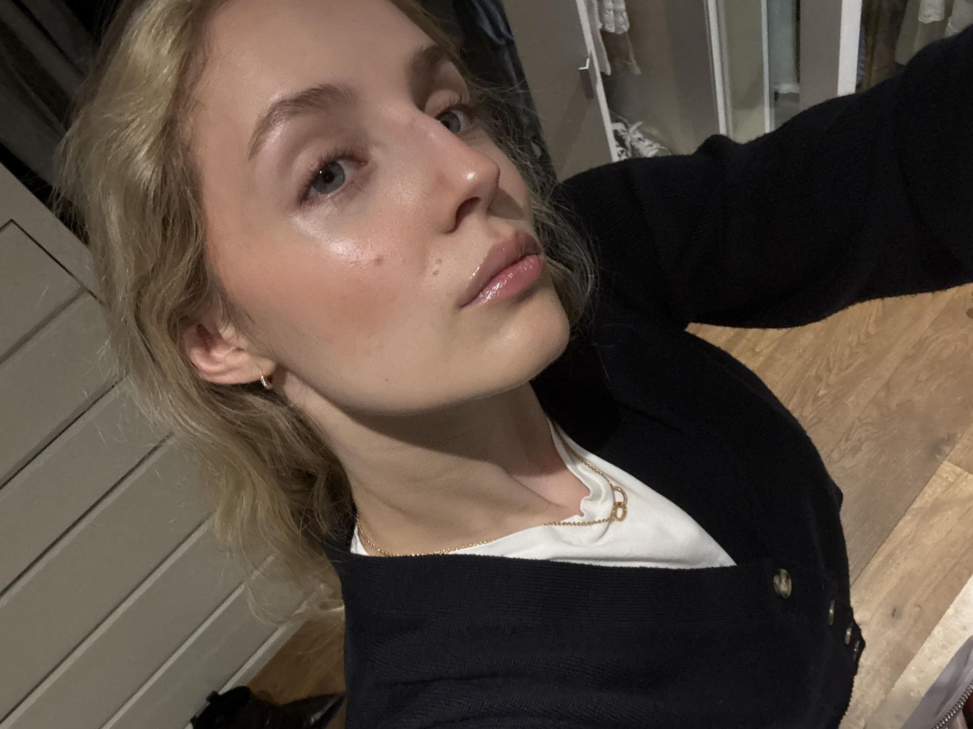 A young Caucasian woman with blonde hair and fair skin is captured in a close-up, high-angle shot. She is wearing a black cardigan over a white t-shirt and a delicate gold necklace with two interlocking rings. Her makeup is subtle, with a dewy finish, and her lips are glossed. The background appears to be indoors, with a slatted wall behind her and wooden flooring beneath. There is no indication of her specific location within Mexico City, Mexico, nor any discernible actions or interactions. The lighting is soft and natural, creating a gentle illumination on her face.