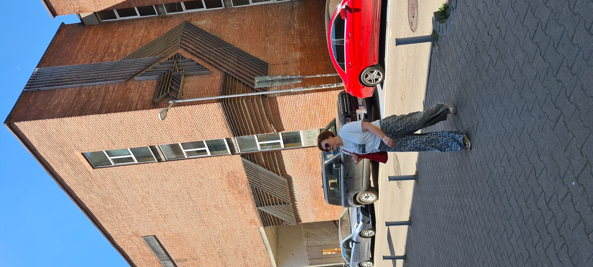 A woman with short brown hair and sunglasses is walking on a sidewalk in front of a modern brick building in Sofia, Bulgaria. She is wearing a white t-shirt with a graphic print and wide-leg patterned pants. She is carrying a red bag over her right shoulder. Behind her, parked on the street, are a silver sedan, a dark gray SUV with the word "DEFENDER" visible on the back, and a red sedan. The building features a distinctive geometric metal facade element. The sky is clear blue.