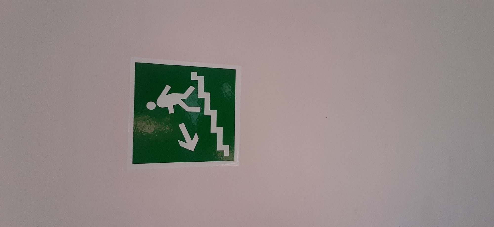 The image shows a green and white safety sign mounted on a plain, light-colored wall. The sign depicts a stylized white figure descending a staircase, with an arrow pointing downwards. This is a standard "down the stairs" or "emergency exit stairs" sign. The figure appears to be in motion, suggesting the act of moving down. The background is a solid, neutral color, providing no additional context about the environment beyond it being an indoor setting with artificial lighting. There is no visible text on the sign or the wall. The image is well-lit, with no indication of time of day or weather conditions. The overall impression is that of a functional safety instruction in a public or commercial building.
