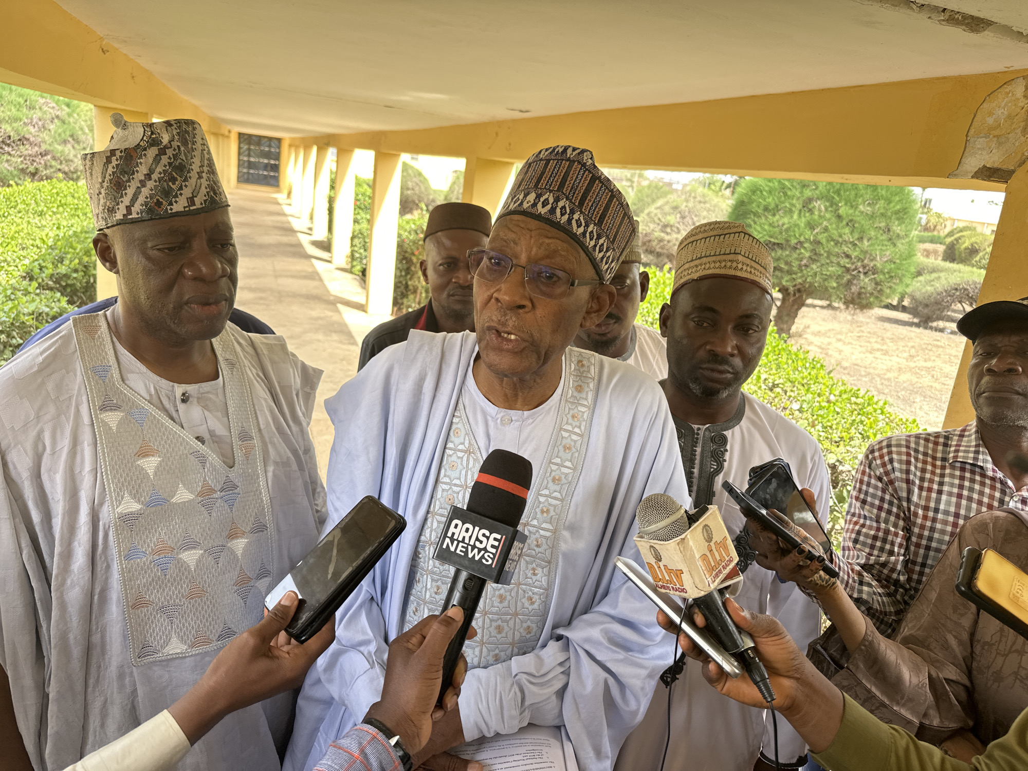 The image depicts a group of men gathered around a central figure who is speaking into several microphones. The location appears to be a covered walkway or veranda, evidenced by the roof structure and exterior landscape visible in the background. The man speaking is wearing a light blue garment with decorative patterns, along with a patterned hat and glasses. He is the focus of attention, as indicated by the array of microphones labeled "ARISE NEWS" and "DITV ALHERI RADIO" held by hands reaching towards him. These hands also hold smartphones, presumably recording the event. Other men are visible in the background, some wearing similar traditional attire. The walkway has tiled flooring, and the outside landscape consists of trimmed shrubs and trees. The event seems to be a press conference or an interview. The city and state are not identifiable in this image.