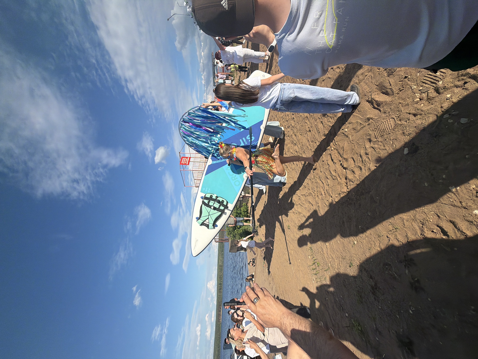 A group of people are gathered on a sandy area near a body of water, likely a lake or river. The sky is blue with scattered white clouds, indicating fair weather and daytime. The scene appears to be outdoors, possibly a beach or a park area.
In the foreground, a person wearing a black baseball cap and a light-colored t-shirt is partially visible, with their arm extended as if holding a camera or phone. The camera is pointed towards a group of people who are the main subjects.
A prominent object in the middle ground is a paddleboard, decorated with blue and silver streamers. A person wearing a grass skirt and a flower lei is standing on the paddleboard, appearing to be part of a performance or a themed event. Another person, with long brown hair, is standing next to the paddleboard, holding onto it and looking towards the person on it. Several other people are scattered around, watching the event or participating in activities. Some individuals are wearing casual clothing, suggesting a relaxed outdoor gathering.
The ground is sandy, with footprints visible. In the background, the water stretches out, and across the water, there is vegetation and possibly some structures. Shadows are cast on the sand, indicating a sunny day. The overall atmosphere seems festive and energetic.
There is some visible text on the paddleboard, but it is mostly illegible. The t-shirt of the person in the foreground has some yellow lettering that appears to say "THE C... RUN".