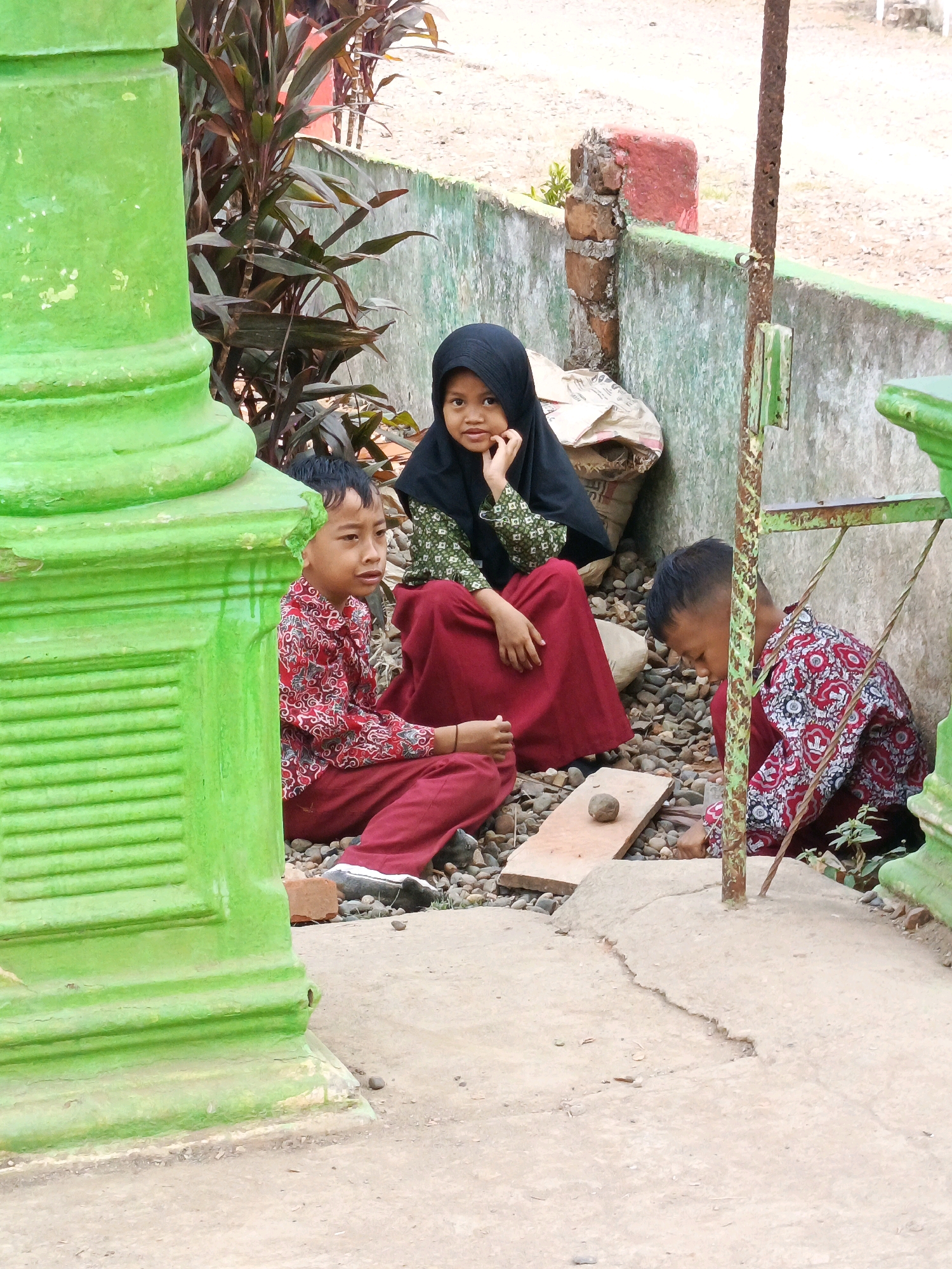 In Megang Sakti IV, Indonesia, three children, two boys and a girl, are sitting on the ground near a green, fluted pillar and a weathered teal wall. The children are dressed in school uniforms, consisting of red and white patterned shirts and red trousers or skirts. The girl, wearing a black hijab and a patterned green top, sits with her elbow on her knee and her chin resting on her hand, looking towards the camera. The boy to her left, also in a red and white shirt and red pants, is looking to the side. The boy to the right, wearing a similar uniform, is hunched over, seemingly engaged in an activity on the ground. In front of them, on a wooden plank, rests a small, round stone. The environment appears to be an outdoor area, possibly a schoolyard, with gravel and cracked concrete on the ground, and some leafy plants visible behind the wall. A rusty metal pole with a green fixture stands near the children.