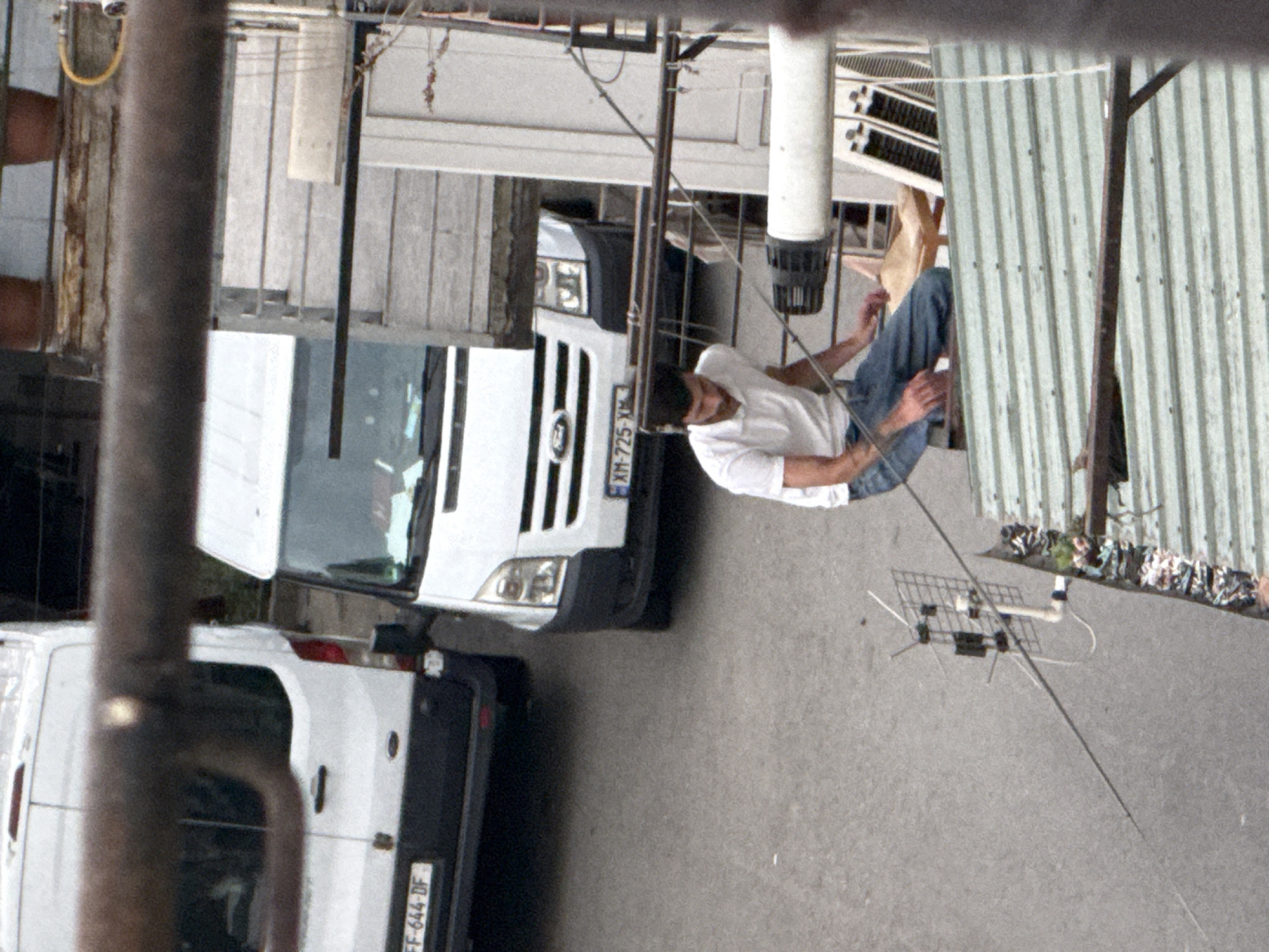 The image shows a man in a white shirt and blue jeans sitting on an elevated platform, possibly a rooftop or balcony, overlooking a street scene. He is leaning back and appears relaxed. Below him, two white vans are parked on the street. One van is closer to the foreground and has its rear doors open, revealing some of its interior. The other van is further back and appears to be a delivery vehicle.
The setting is urban, with corrugated metal structures and buildings visible in the background. A metal pipe or pole runs vertically in the foreground, partially obscuring the view. There is a small, wire antenna-like object on the ground in the middle distance. The lighting suggests it could be daytime, possibly late morning or afternoon, with a slight overcast sky. There is no discernible weather information.
Visible text on one of the vans reads "XM-725-XM" and another van has "FF-644-DF". The overall impression is of a casual, everyday moment in an urban environment. The man's posture and expression suggest a moment of rest or observation. The location context of Tbilisi, Georgia, adds a specific geographical element to the scene.