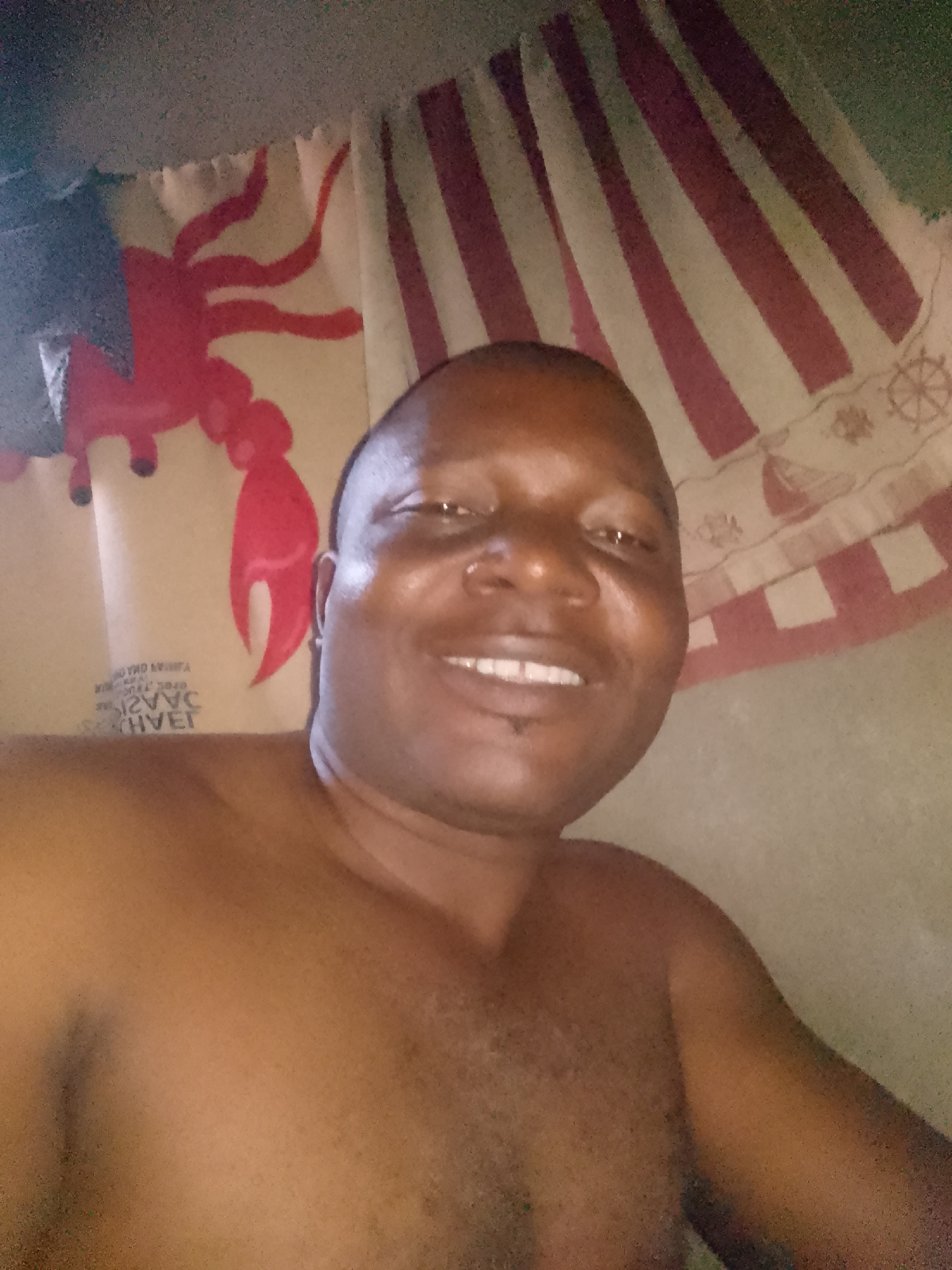 A smiling, dark-skinned man is centered in a close-up shot, shirtless. The background features fabric adorned with images; to the left, a red lobster-like shape is visible, while the right side displays red and white stripes, along with patterns of ships' wheels. The man's location is Unknown, Unknown.