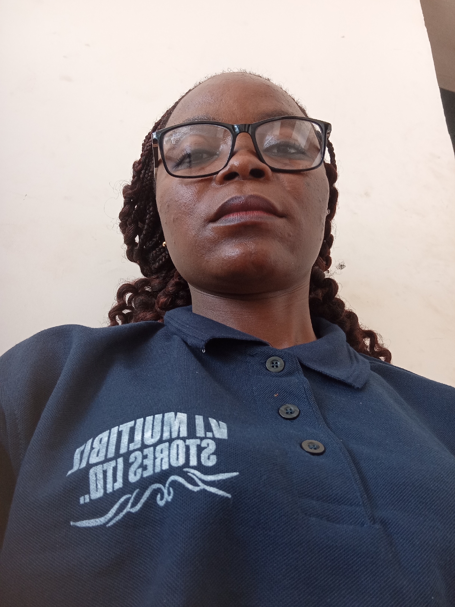 A low-angle, eye-level, medium shot shows a Black woman wearing glasses and a dark blue polo shirt with a white logo. Her braided hair is visible to the sides. The woman is looking directly at the camera with a neutral expression. She has dark skin and is wearing dark-rimmed glasses. The polo shirt has a collar and two black buttons. The white logo on the shirt is partially obscured and appears to be text and a decorative flourish. The background is a plain, light-colored wall. The scene appears to be indoors. The woman is wearing small gold earrings. There is a faint sheen of sweat on her forehead. The logo on the shirt reads "JALITIUM I.V. STORES LTD" in white text. The image was taken in Jalingo, Nigeria.