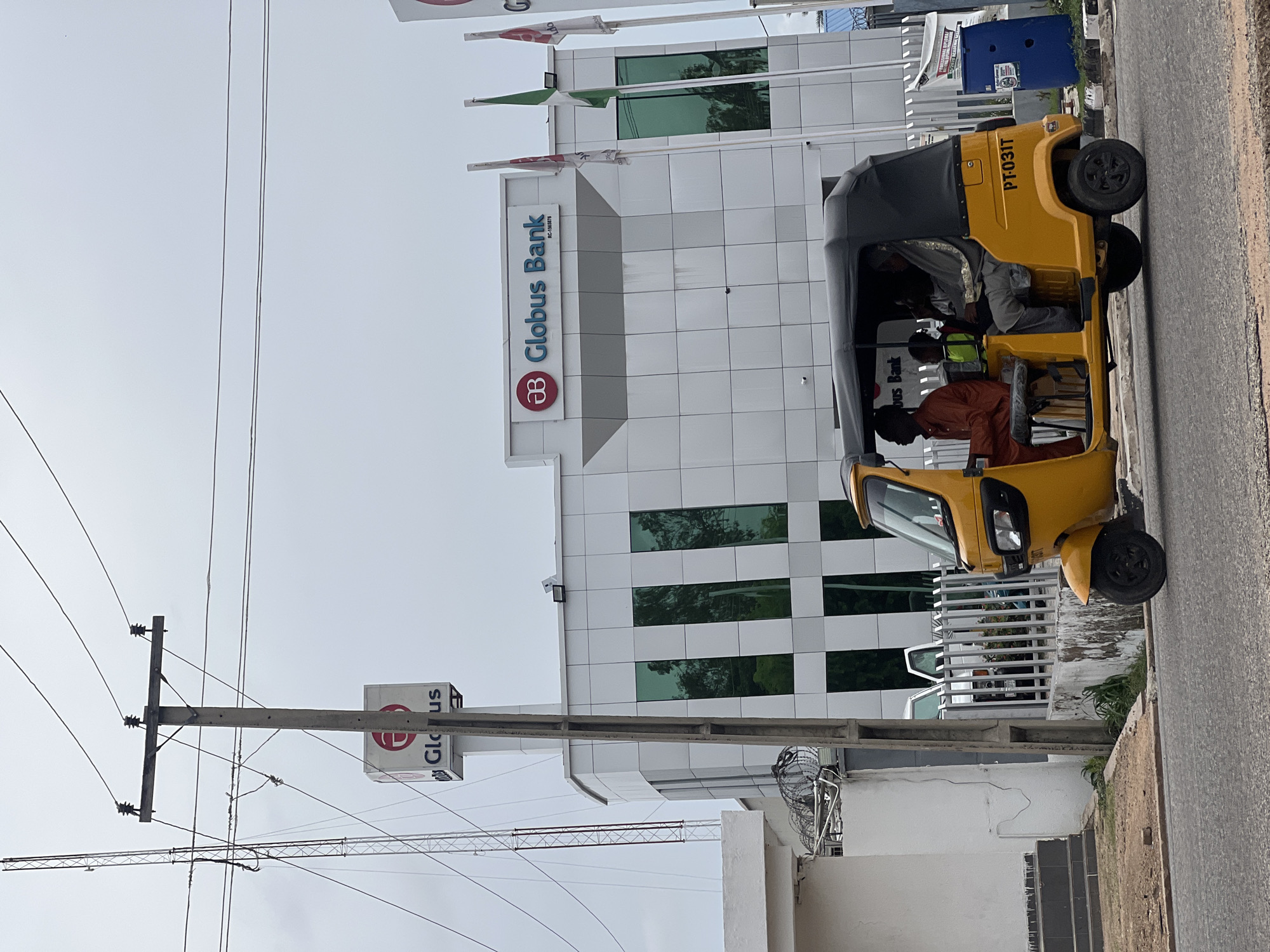 A yellow auto-rickshaw with a black canopy is moving along a paved road in front of a modern, white building with a patterned facade and reflective green windows. The auto-rickshaw is carrying three passengers; one driver in an orange outfit, and two passengers in the rear, one in grey clothing and another in a green vest. The license plate of the auto-rickshaw reads "PT-031T." Above the auto-rickshaw, on the building, the Globus Bank logo and name are visible. A tall, concrete utility pole with wires runs diagonally across the left side of the image. In the background, a lattice tower structure is visible. The sky is overcast. This scene is located in Jalingo, Nigeria.