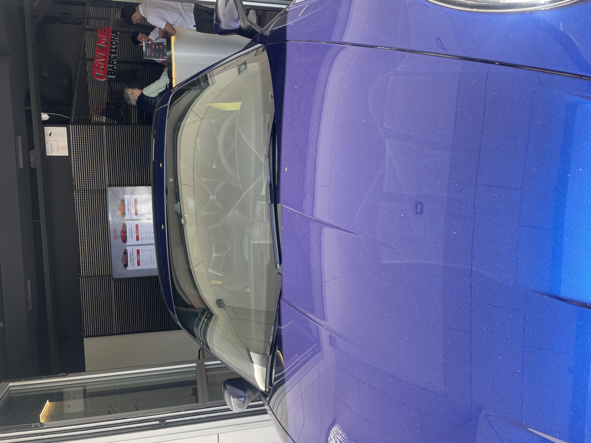 The image captures an indoor scene, dominated by a vibrant blue (likely metallic purple-blue) sports car positioned in the foreground. The car's windshield reflects its interior, including what appears to be a dashboard with "GT3" visible in reverse, suggesting a Porsche GT3 model. The car's paintwork shows visible dust or small specks.
In the background, a reception area is visible. Three individuals are present at a counter: two men stand behind it, one in a white t-shirt and the other in a light-colored shirt, seemingly assisting an older man with grey hair wearing a light green shirt and dark vest, who stands in front of the counter. They appear to be engaged in a transaction or discussion.
Behind the counter, a prominent red sign reads "DRIVEME BARCELONA", identifying the establishment as a car experience or rental service in Barcelona. To the right of the counter, display posters feature images of luxury cars with associated pricing. One poster clearly shows a red car, labeled "FERRARI 458 SPECIALE," with a price of "555 €." The setting features dark, possibly slatted walls and bright indoor lighting, with a large window or glass partition visible on the left. The scene depicts activity at a luxury car experience venue.